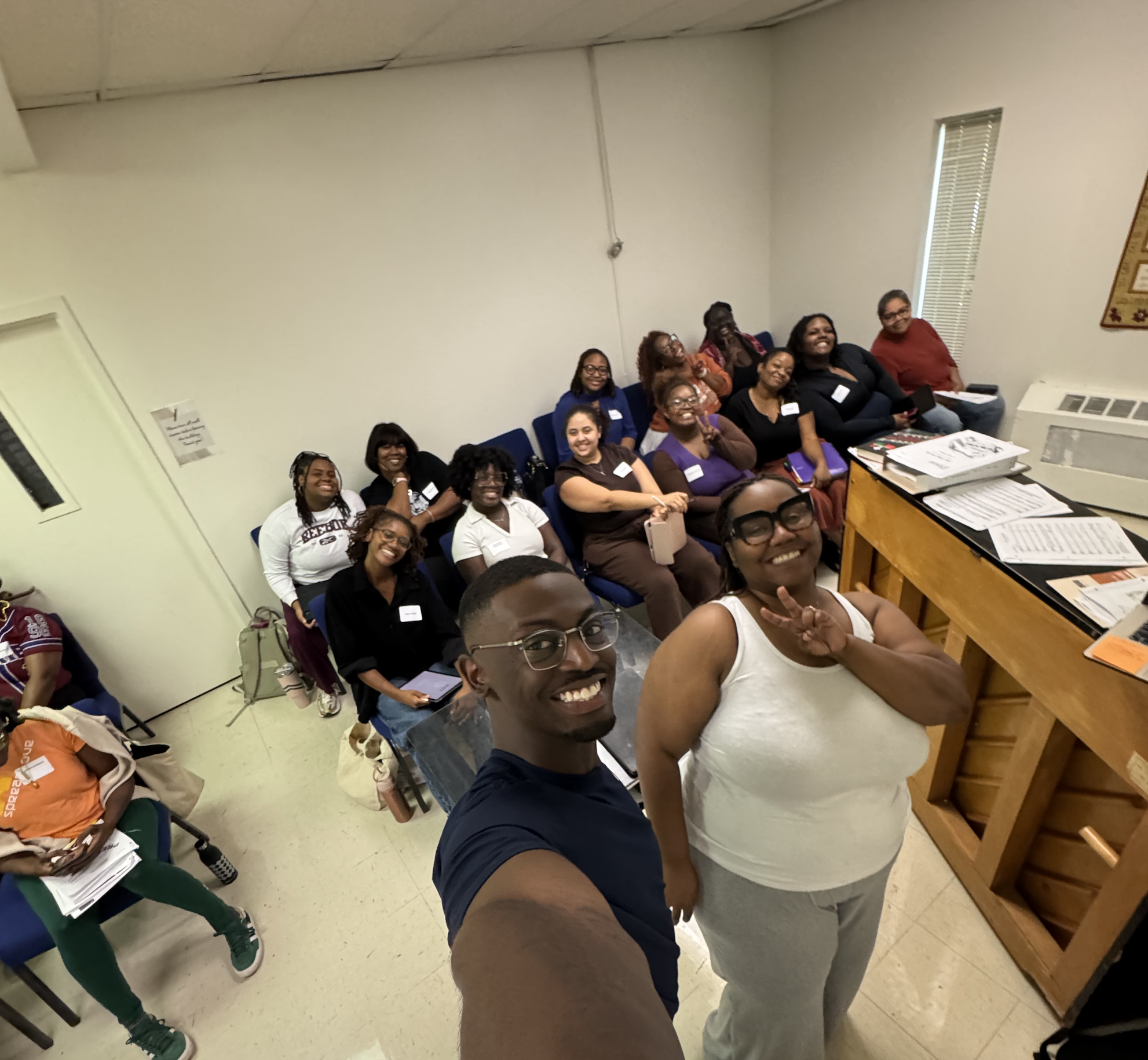 Group of diverse women smiling and taking a selfie in a classroom or meeting room with papers and books on the table.