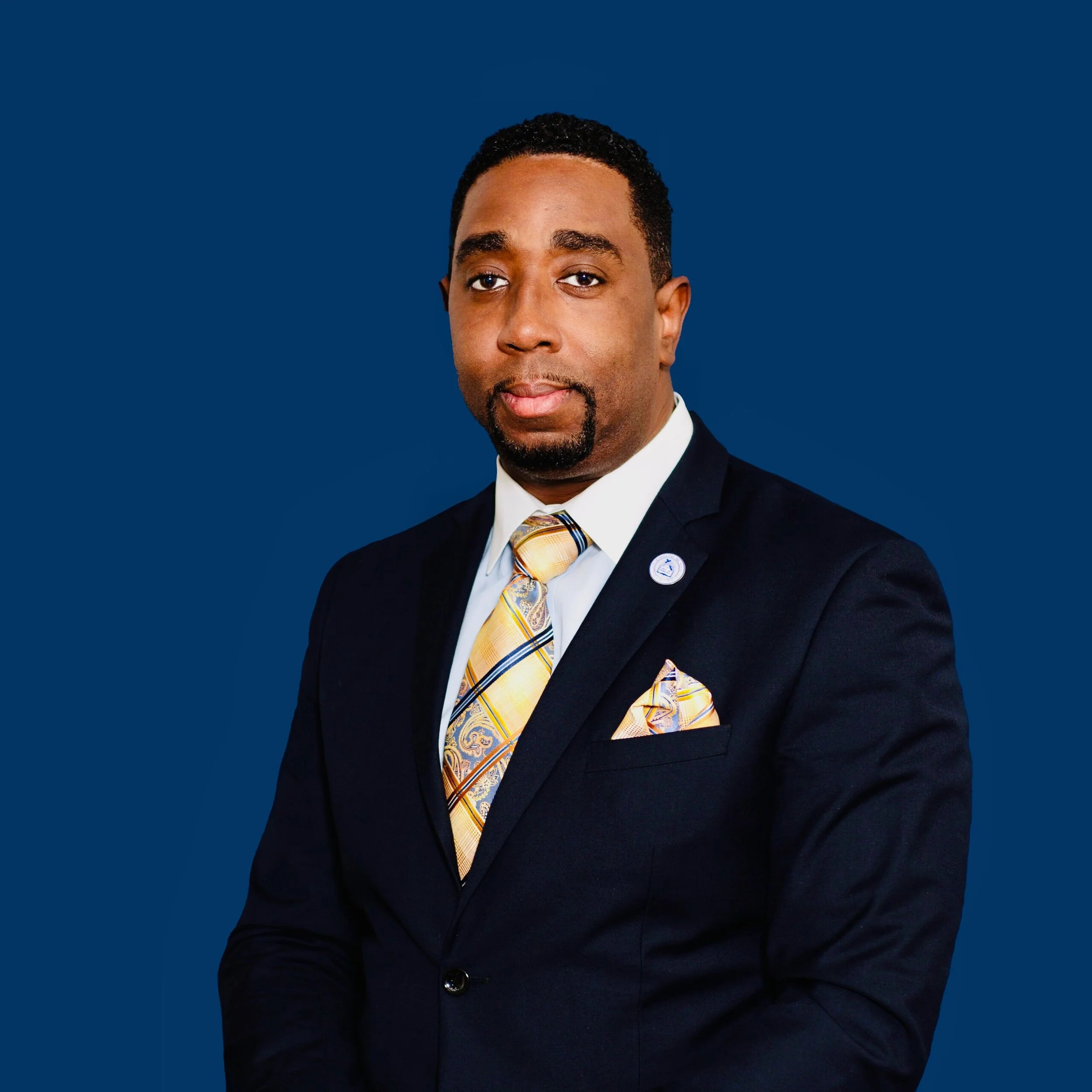 Close-up of a man in a navy suit, white shirt, and yellow plaid tie against a solid dark blue background.