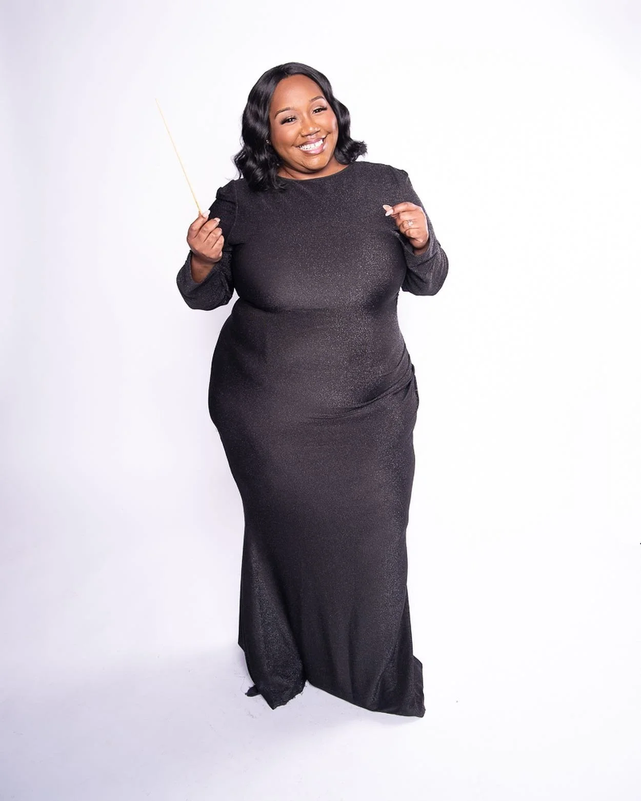 A smiling woman wearing a long black, sparkly dress, holding a conductor's baton, posing against a plain white background.
