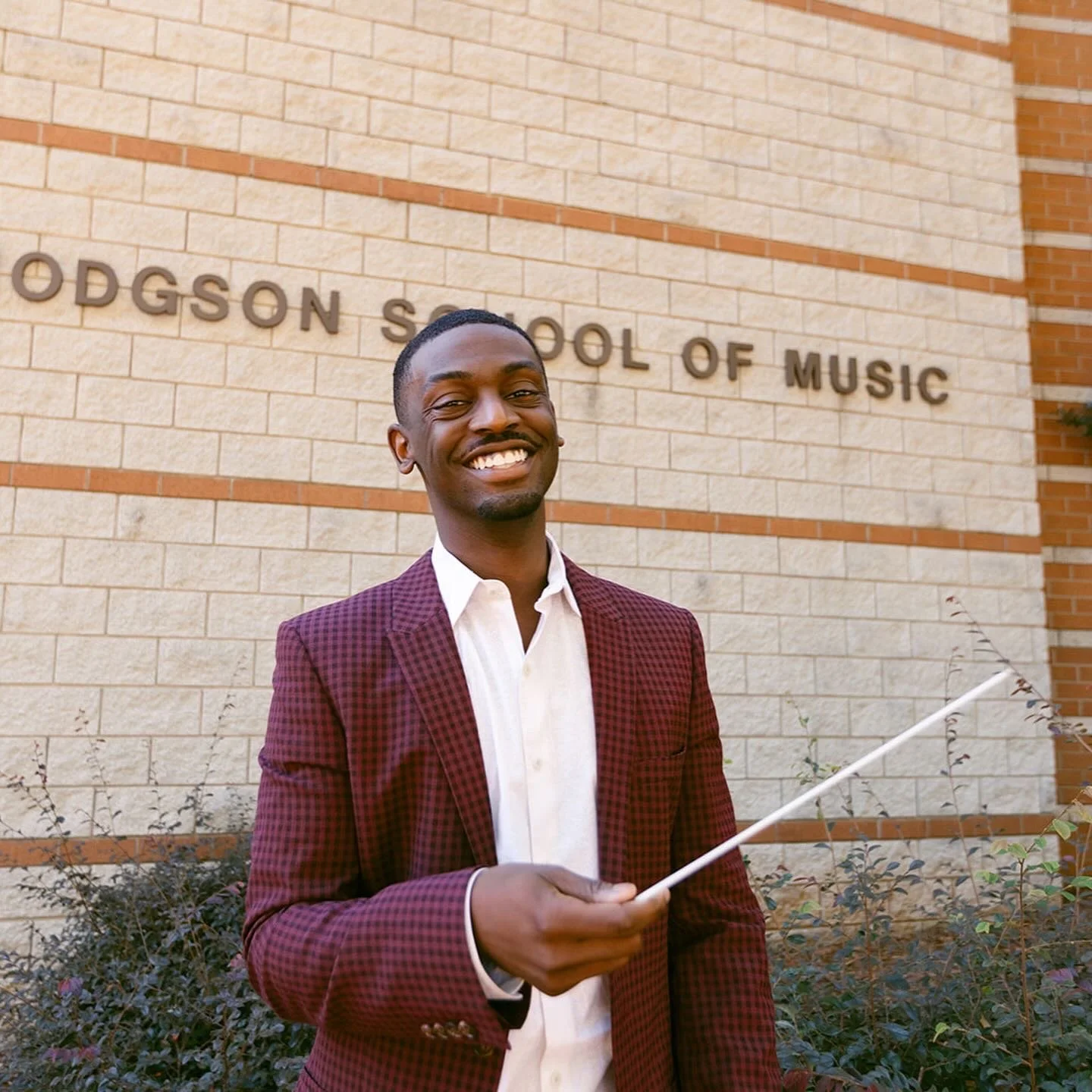 A smiling man in a burgundy blazer and white shirt holding a conductor's baton stands outside in front of a beige brick wall with the words "ODGSON SCHOOL OF MUSIC".