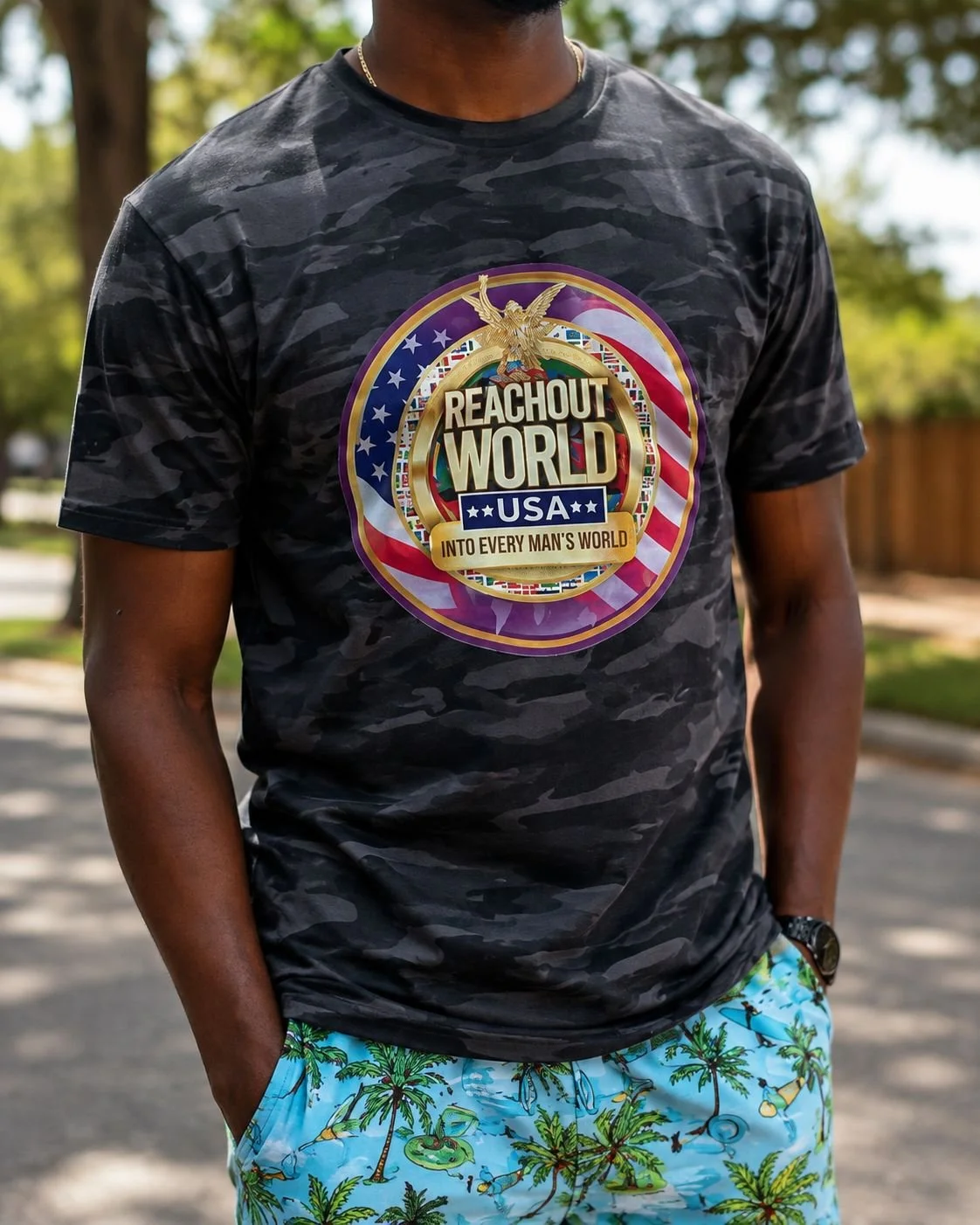 A person wearing a black camouflage T-shirt with a colorful logo that says "Reach Out World USA" and "Into Every Man's World." The person is also wearing blue shorts with a tropical palm tree pattern and has a watch on their left wrist.