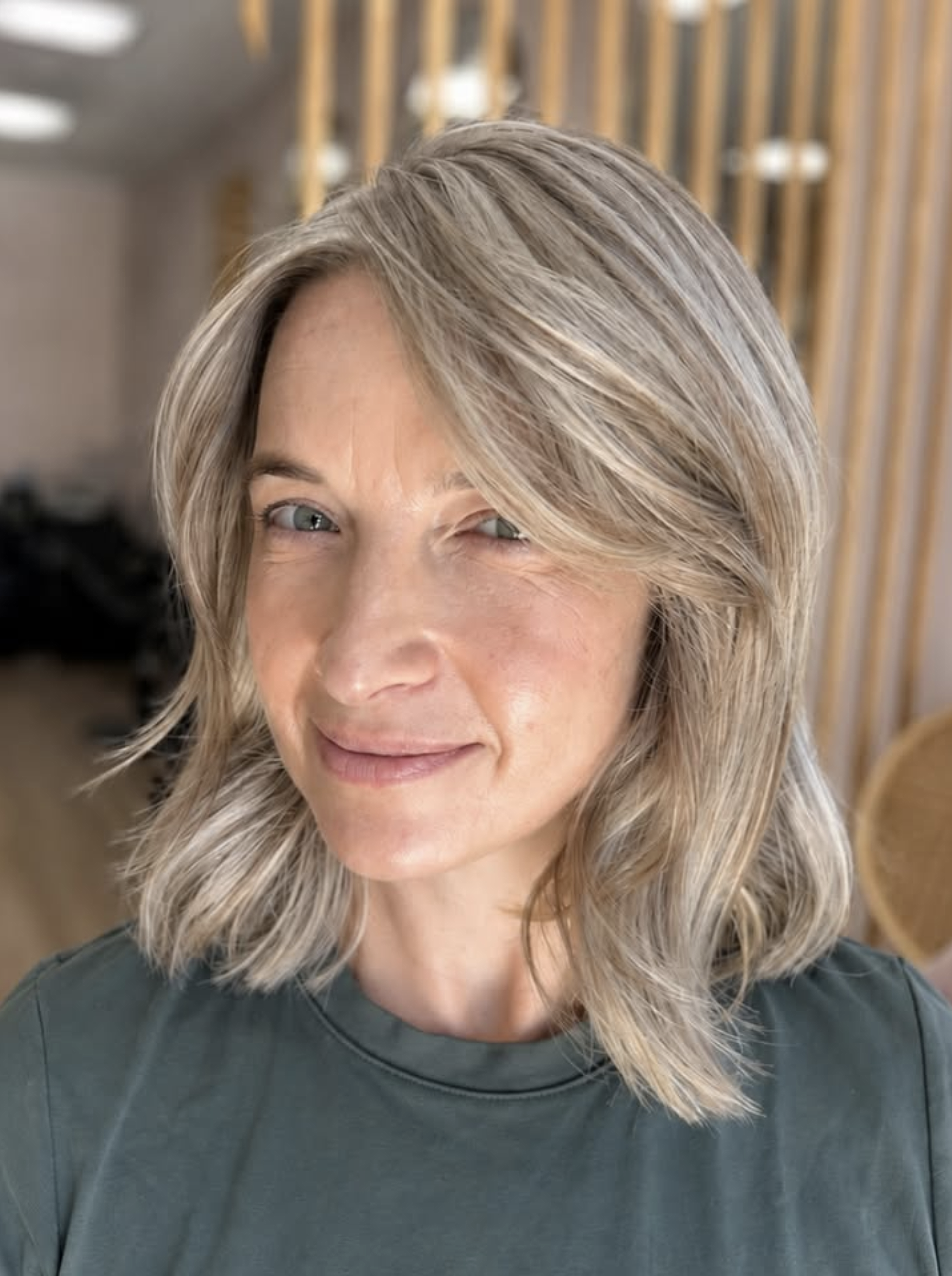 A woman with shoulder-length, wavy blonde hair and light skin, smiling in a black top, in front of a wooden slat background.
