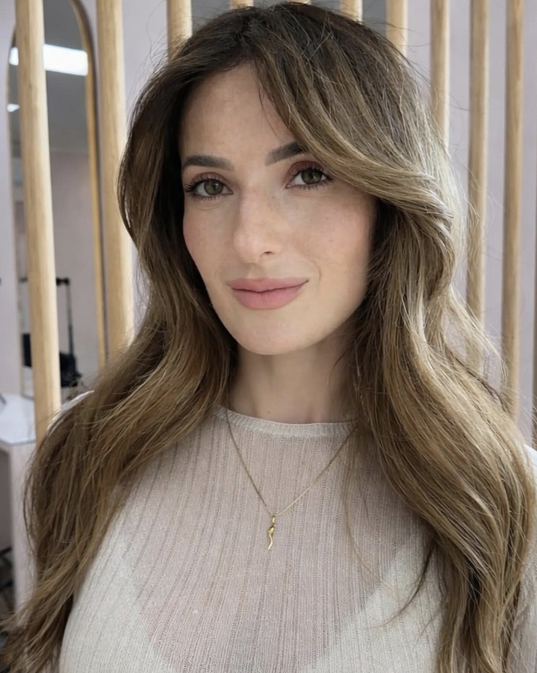 Close-up of a woman with long, wavy brown hair and light skin, standing indoors in front of a background with wooden slats and blurred elements.