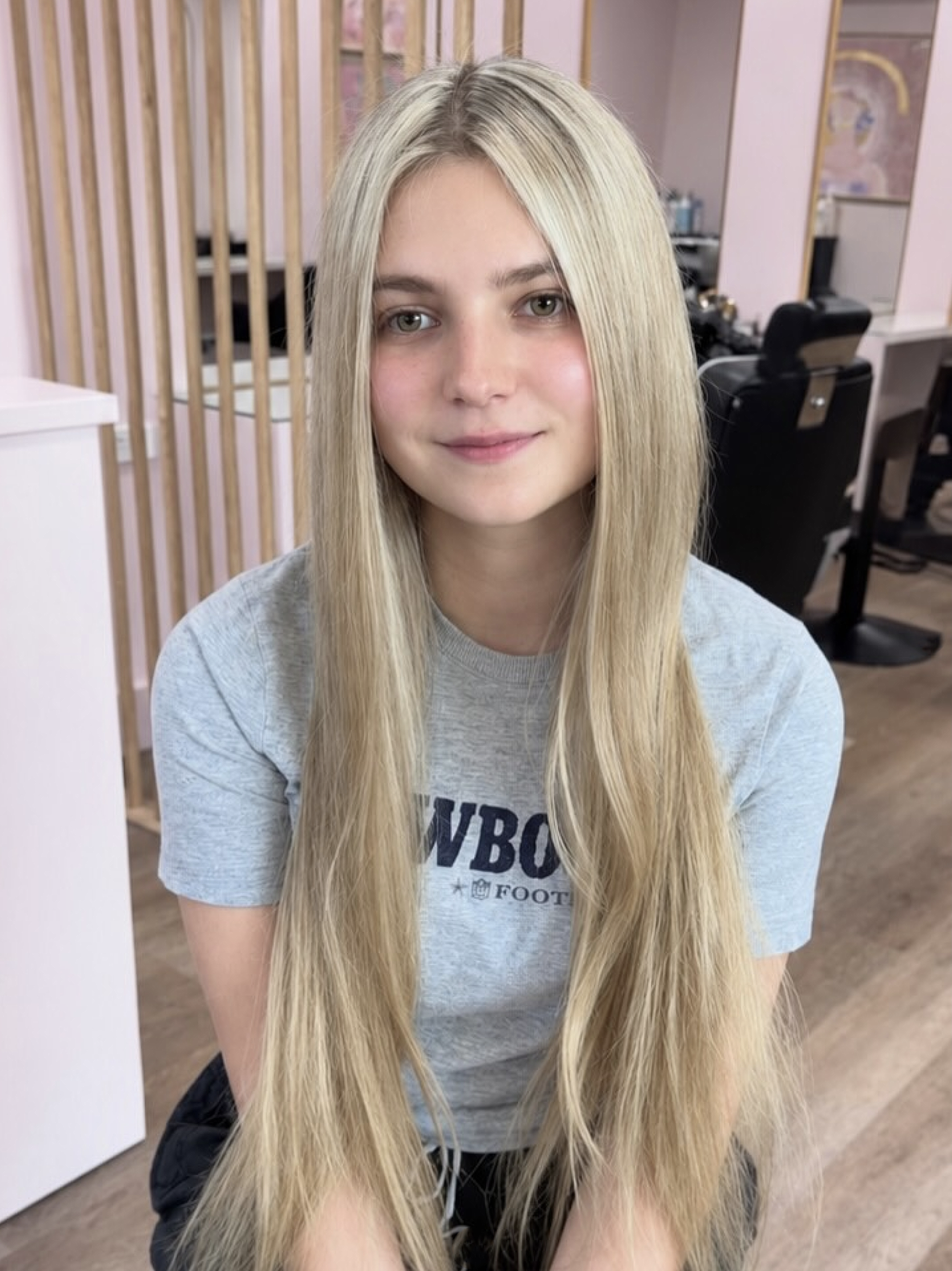 A young woman with long blonde hair and light skin sitting in a hair salon, smiling softly and looking at the camera.