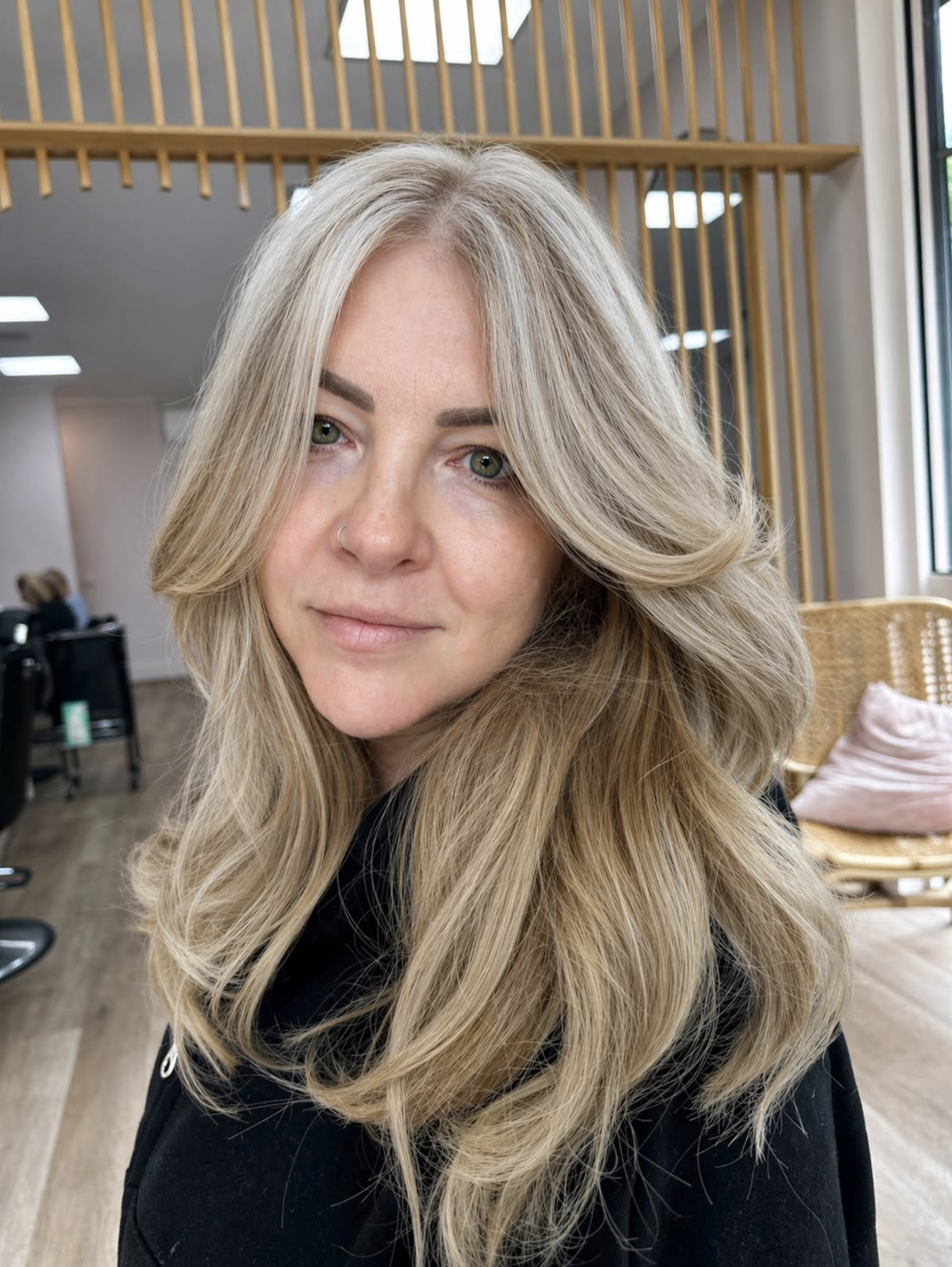 Close-up of a woman with long, wavy blonde hair and green eyes, sitting indoors with wooden architectural elements and large windows in the background.