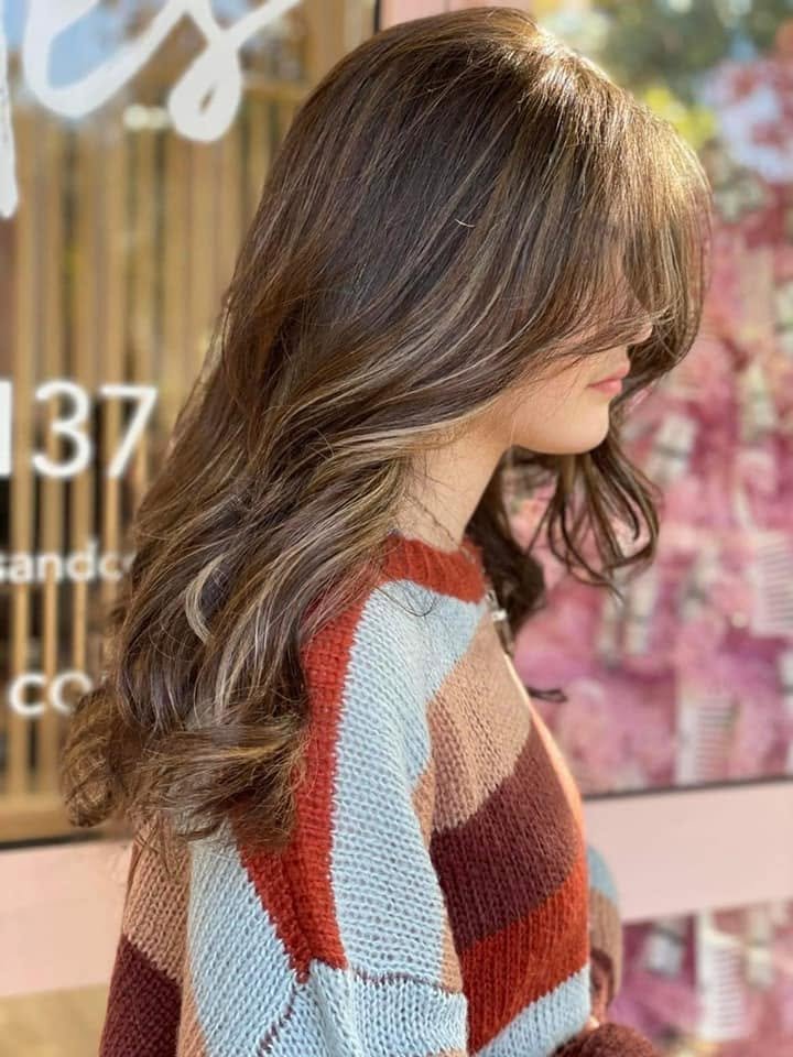 A woman with long, wavy brown hair standing in a store or salon, wearing a colorful striped sweater.