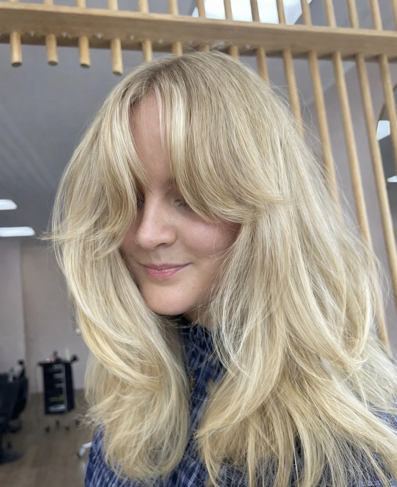 A woman with long, blonde, wavy hair and fair skin is looking down and smiling softly in a salon or studio setting.