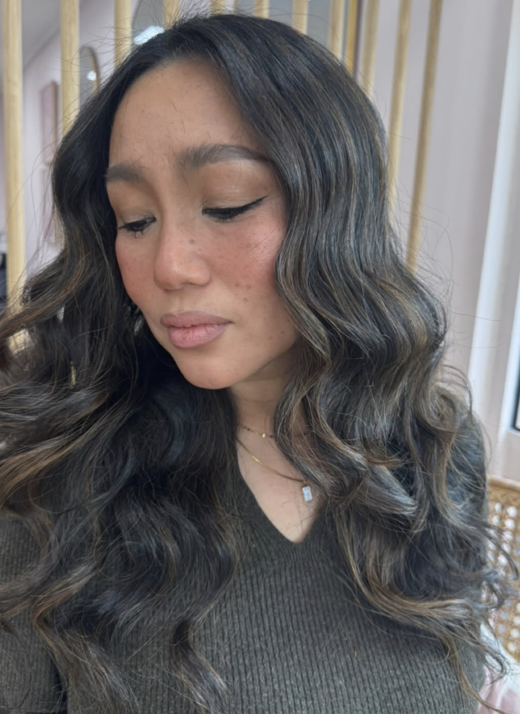 Close-up of a woman with long, wavy black hair with caramel highlights, wearing a dark top and a gold necklace, in a setting with wooden decor in the background.