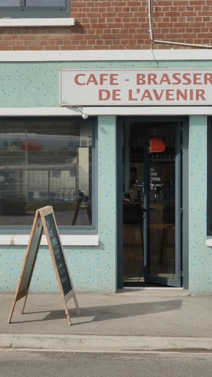 Façade d'un café-brasserie nommé 'De l'Avenir', avec une enseigne en lettres rouges, une porte d'entrée en verre et un tableau noir sur un support en bois à l'extérieur