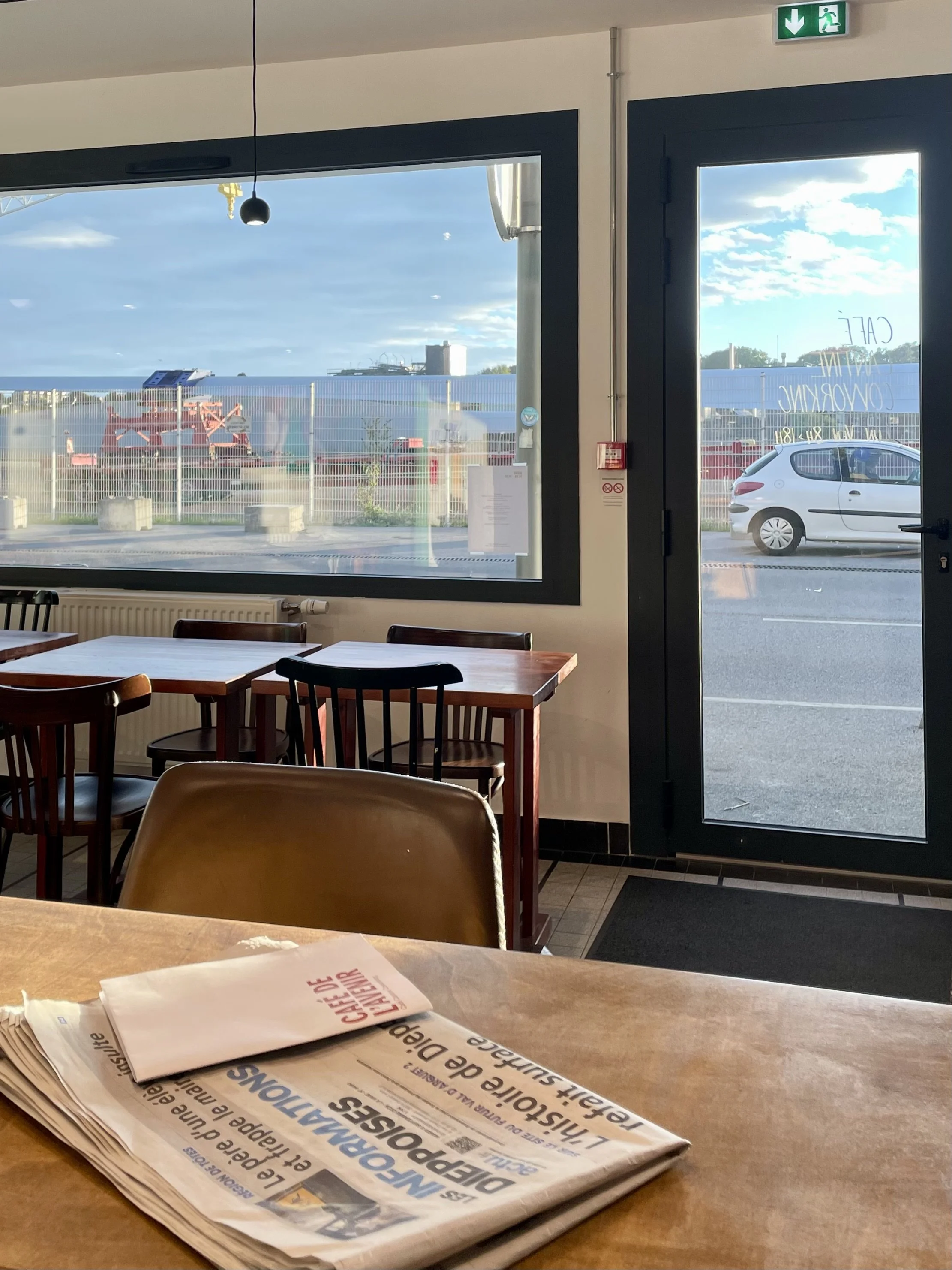 Une table en bois dans un café avec des journaux pliés. On voit des fenêtres donnant sur une rue avec une voiture blanche garée, un panneau de sortie de secours vert, et un ciel ensoleillé avec des nuages.