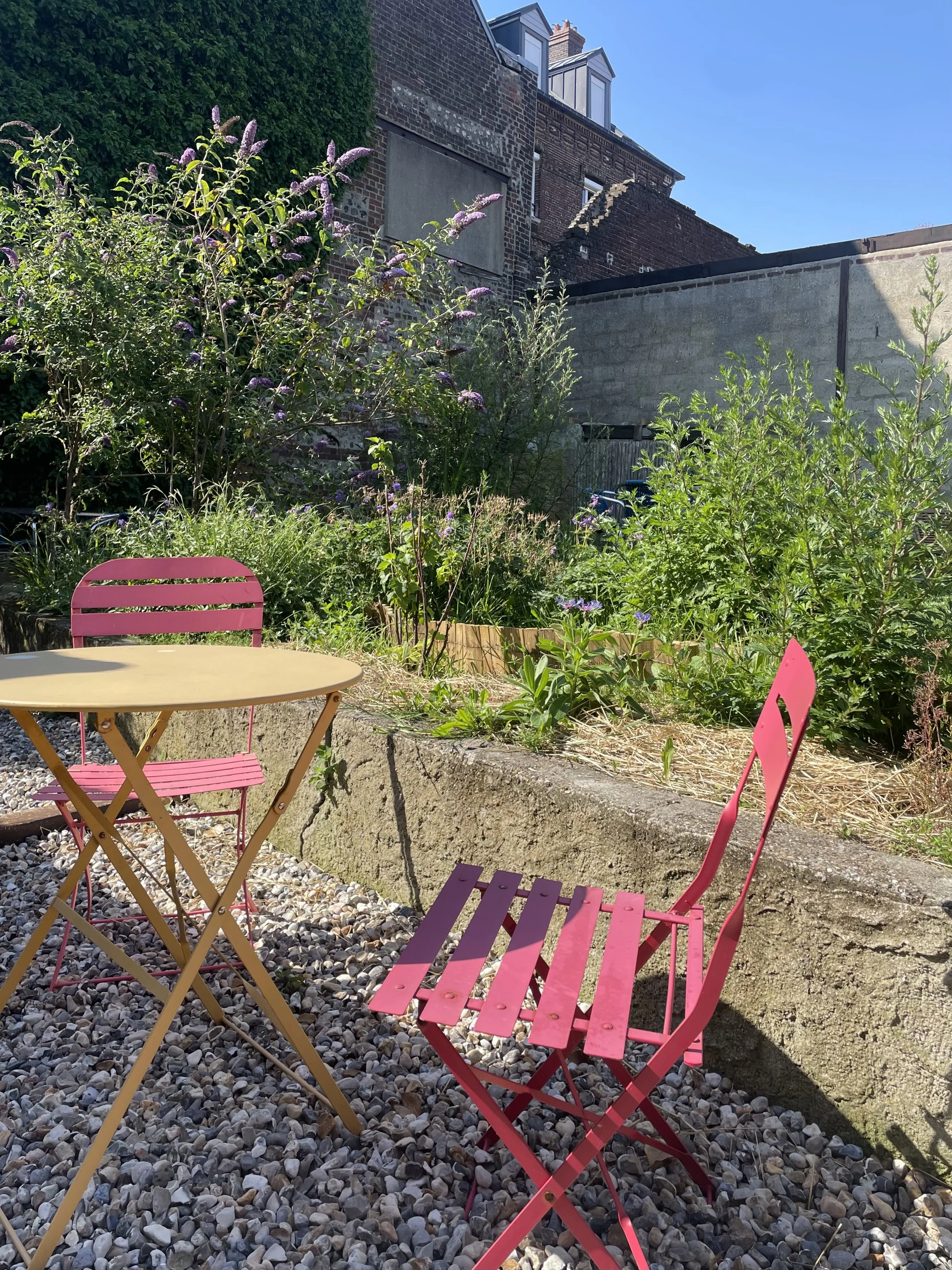 Une petite terrasse en plein air avec une table jaune et des chaises roses et fuschia, entourée de végétation luxuriante et de pavés, avec un bâtiment en brique en arrière-plan sous un ciel bleu clair.