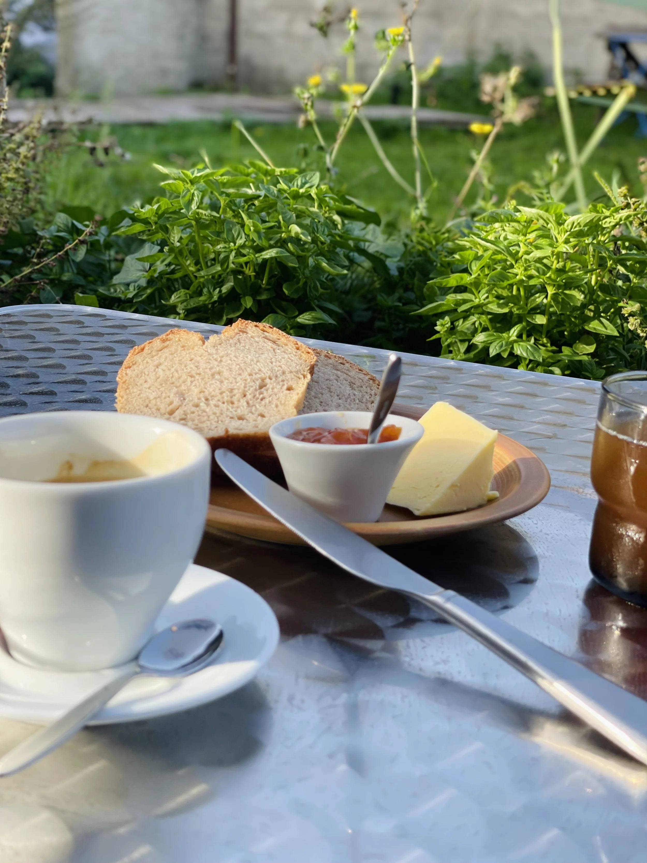Petit déjeuner avec du pain, du beurre, de la confiture, du fromage, des œufs et un verre de boisson, servi sur une table à l'extérieur, avec un fond de végétation verte.