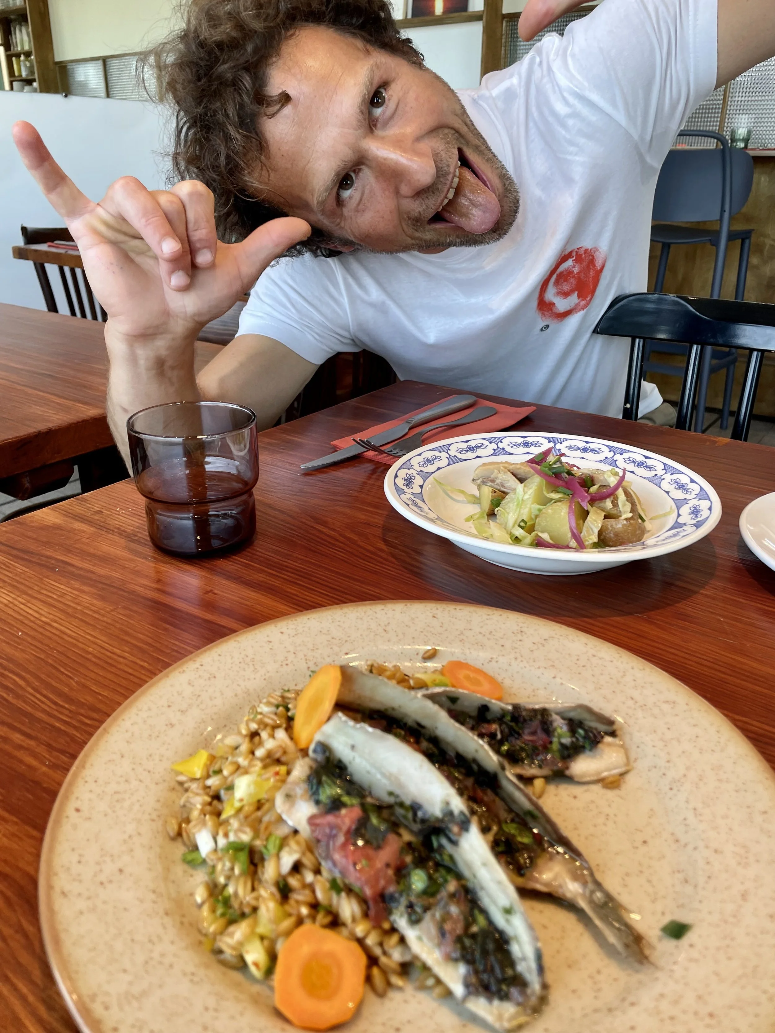Un homme avec des cheveux bouclés, fait un geste de paix avec la main, souriant, avec une langue sortie, assis à une table avec un plat de poisson, légumes et céréales, dans un restaurant.