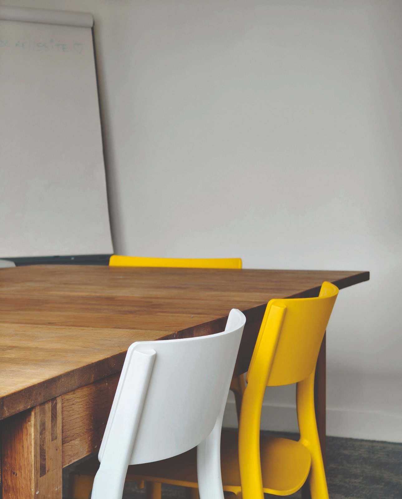 Une table en bois avec deux chaises, une blanche et une jaune, dans une pièce avec un mur blanc et un tableau blanc.