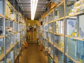 A narrow aisle in a warehouse with shelves stocked with boxes and packages on both sides.