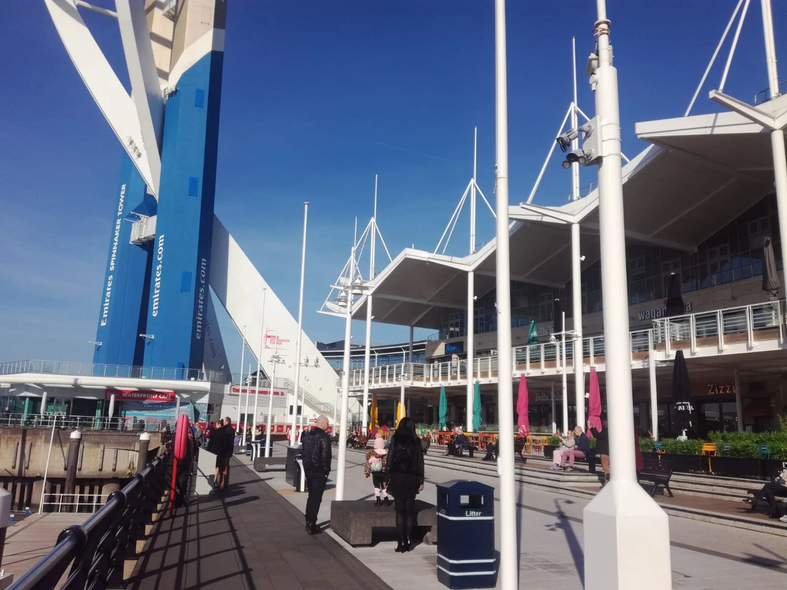 People walking along a promenade outside a large, modern stadium with a blue and white exterior and a prominent Emirates Spinnaker Tower in the background.