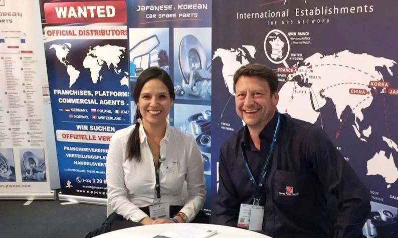 Two people, a woman and a man, sitting at a table with trade show badges, smiling in front of a display booth with maps and promotional posters related to Japan-Korea car spare parts export and international automotive distributors.