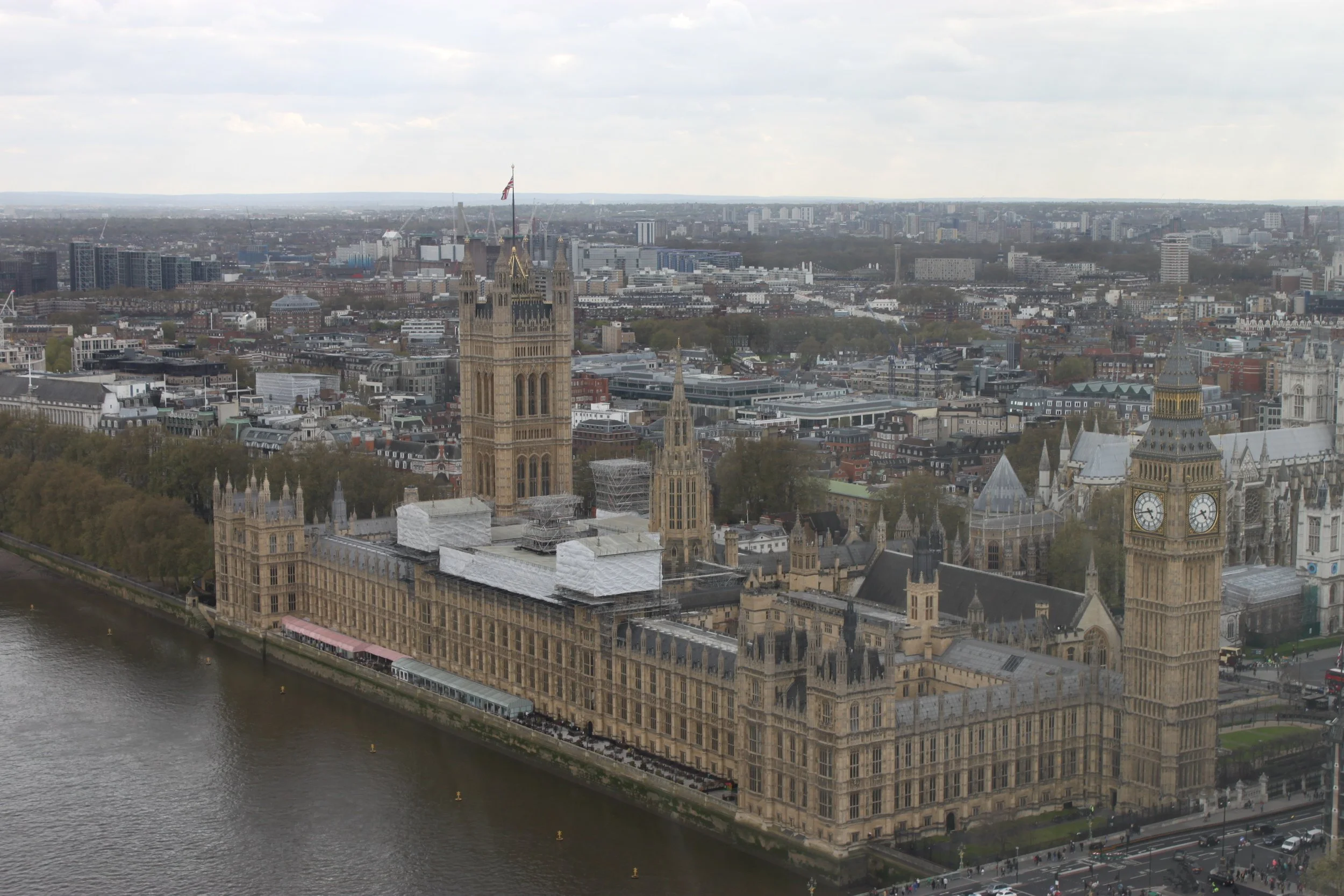 A panoramic view of London's skyline featuring the historic Palace of Westminster with Big Ben clock tower, situated along the River Thames with boats and a riverbank walkway. Modern buildings extend into the cityscape under a cloudy sky.