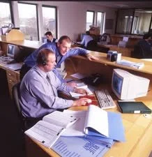 Four men working at desks in an office, surrounded by papers, files, and computers.