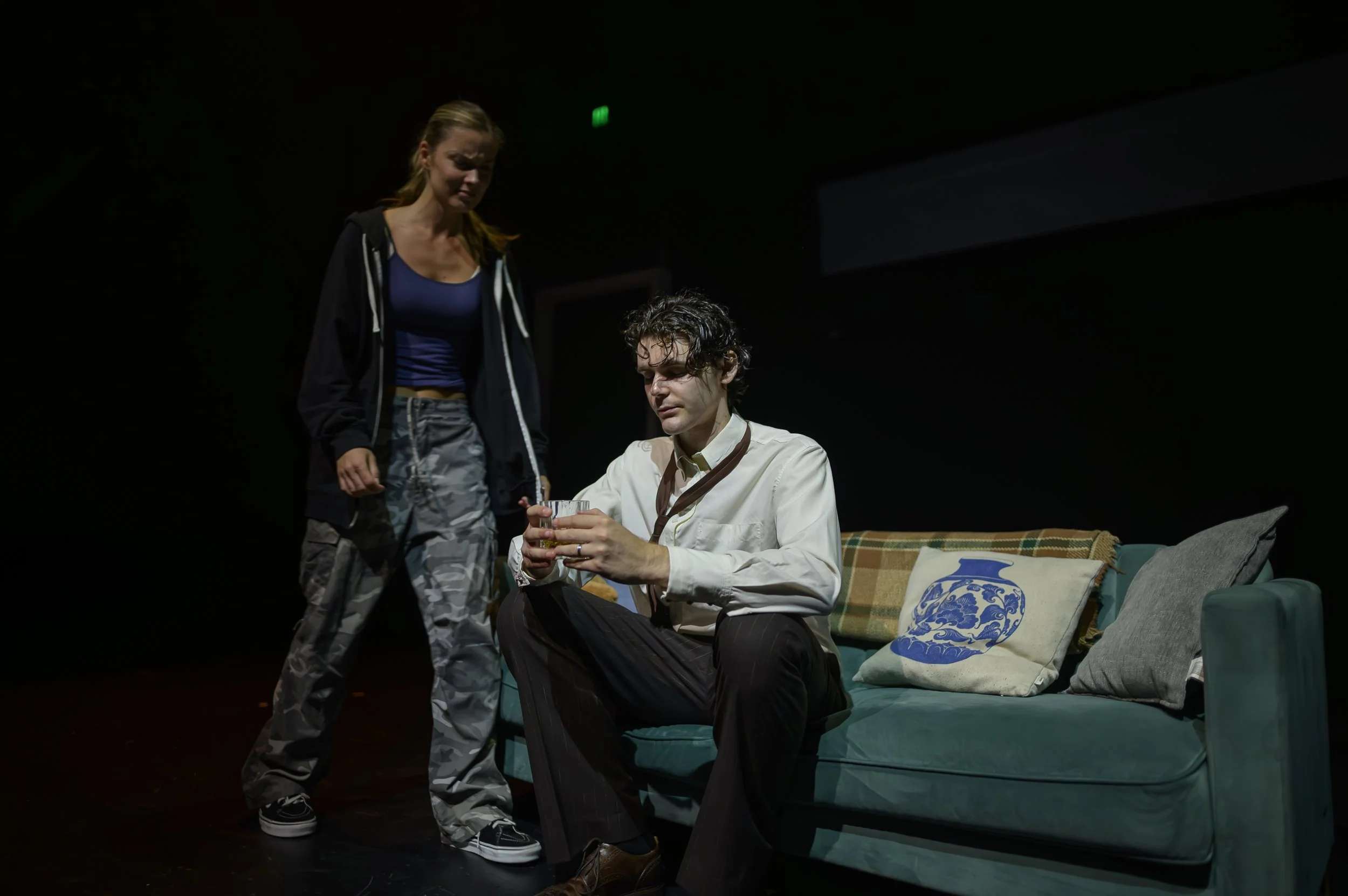 Two performers engaged in a dramatic scene, with one sitting on a green couch holding a glass of whiskey and the other standing close by in a dark room.