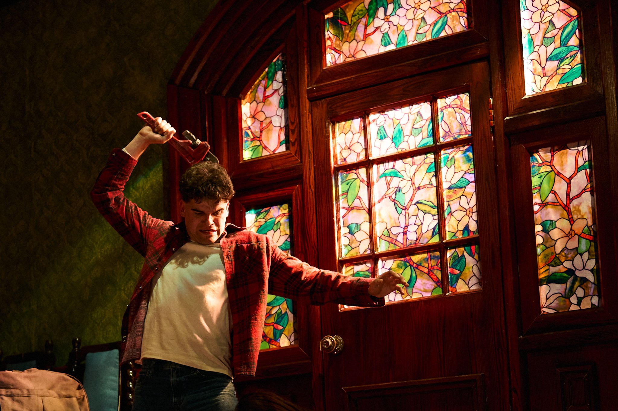 A person with curly hair wearing a red plaid shirt and a white shirt performs on stage, holding a wrench, in front of stained glass windows with floral patterns.