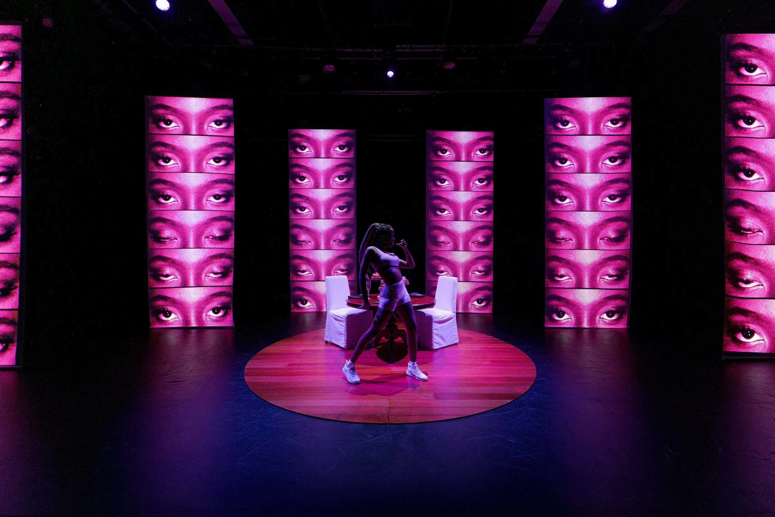 A person performing on stage in front of multiple large screens displaying close-up images of a woman's eyes and eyebrows, with vibrant purple lighting.