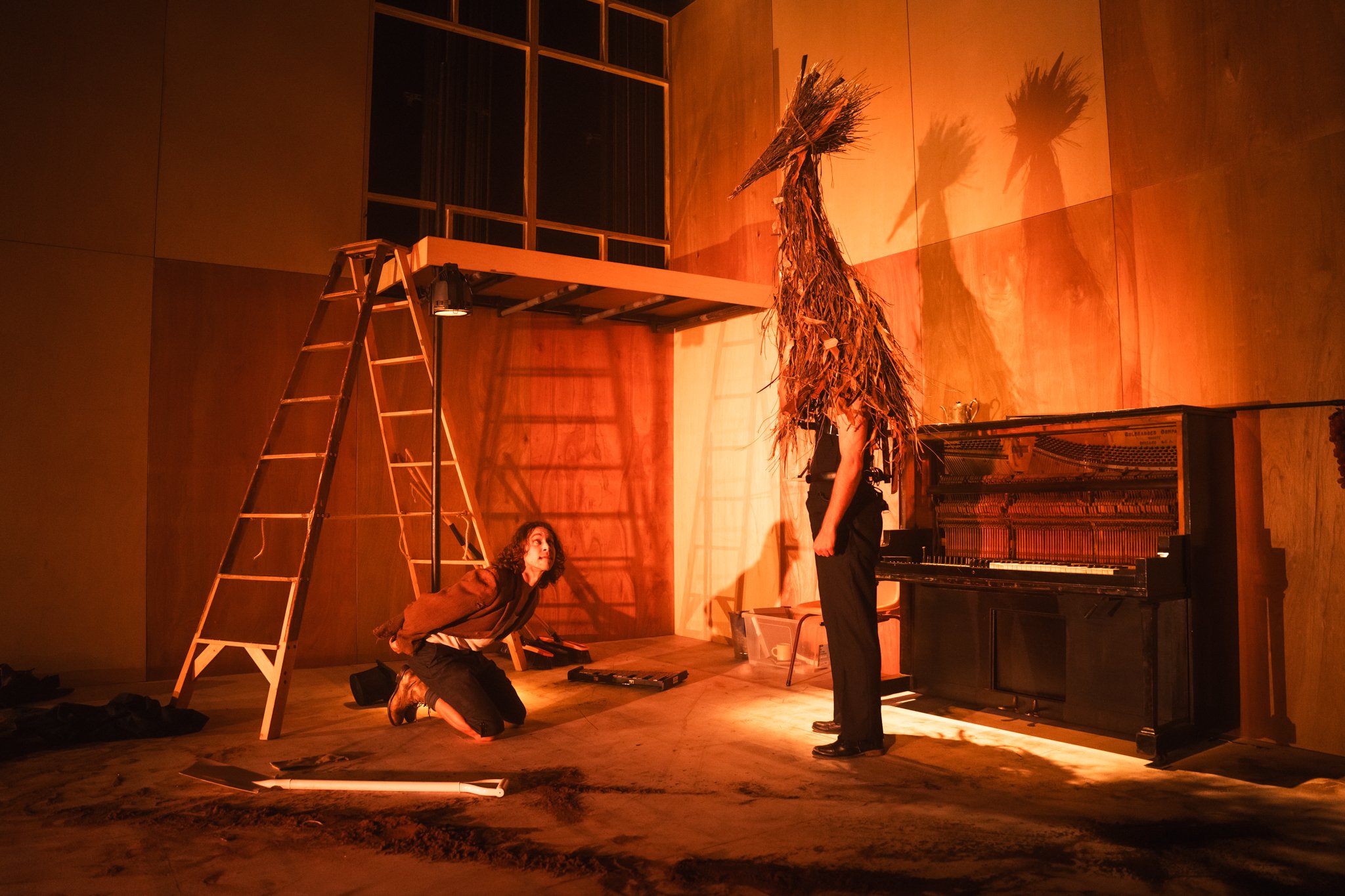 Two people performing on a stage with a wooden platform, ladder, and a bird costume made from a bundled arrangement of dried branches, illuminated by warm orange lighting in a wooden-paneled room.