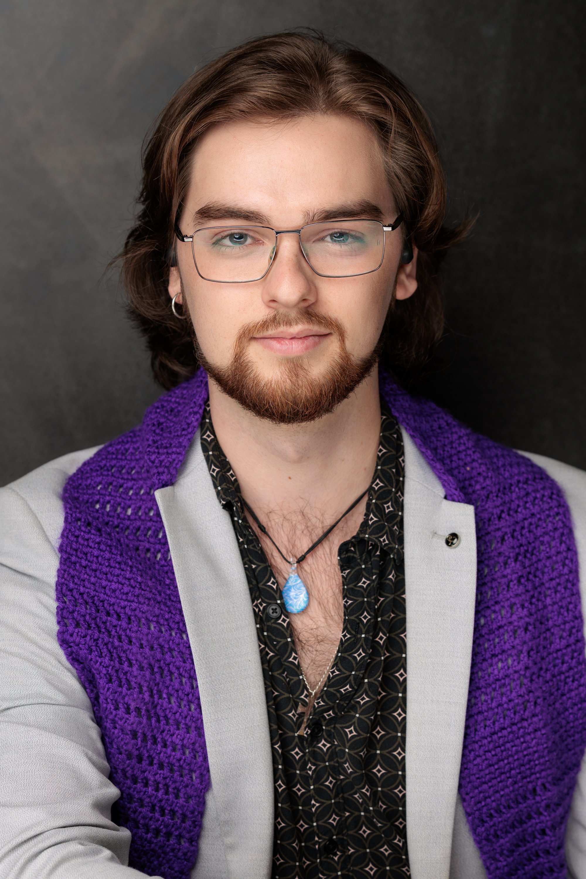 A young man with shoulder-length brown hair, glasses, a beard, and mustache, wearing a purple knitted scarf, a patterned black shirt, and a light gray blazer, standing against a dark background.