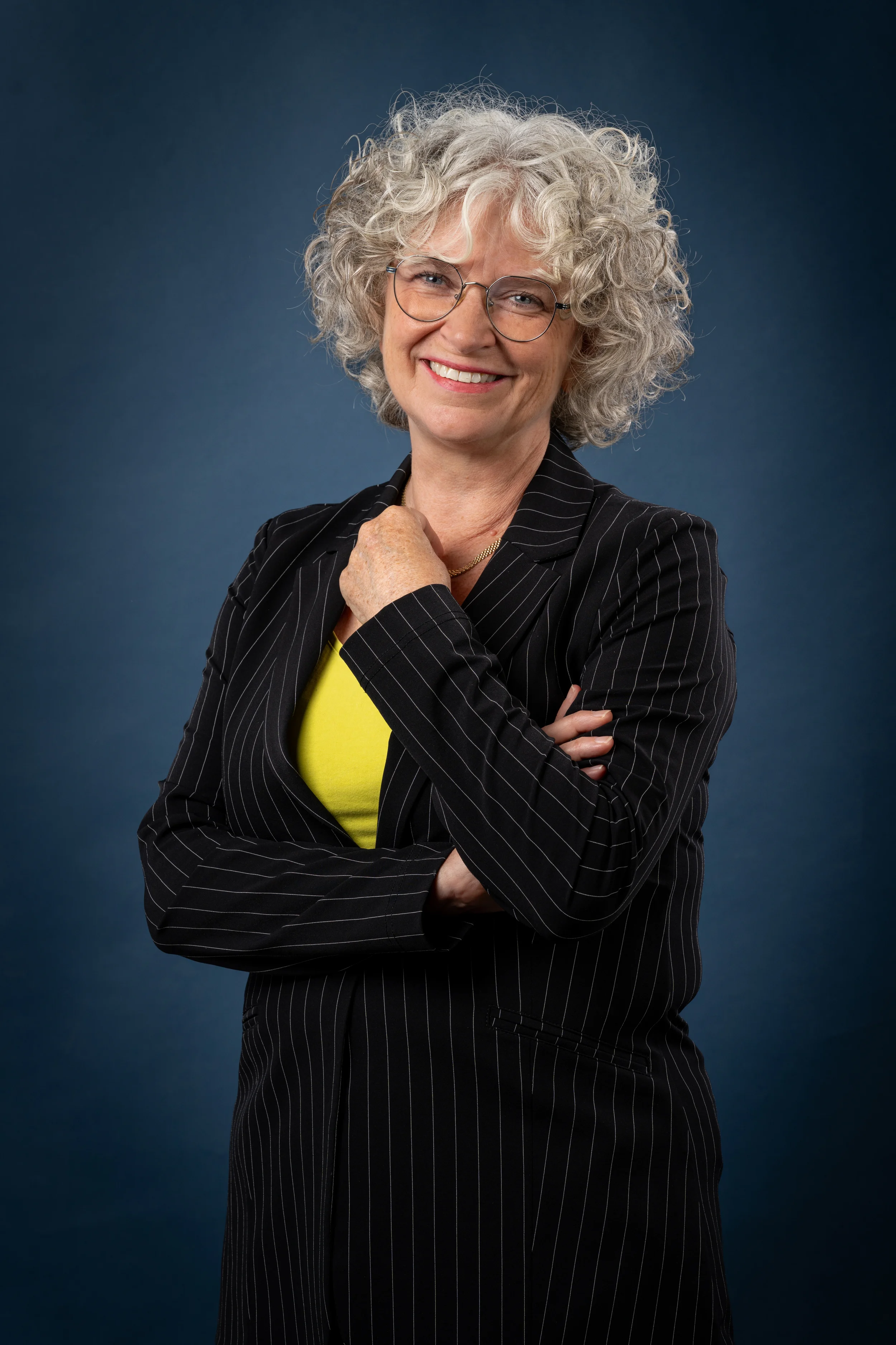 A woman with curly silver hair, wearing glasses, a black pinstripe blazer, and a bright yellow top, smiling with her arms crossed against a dark blue background.