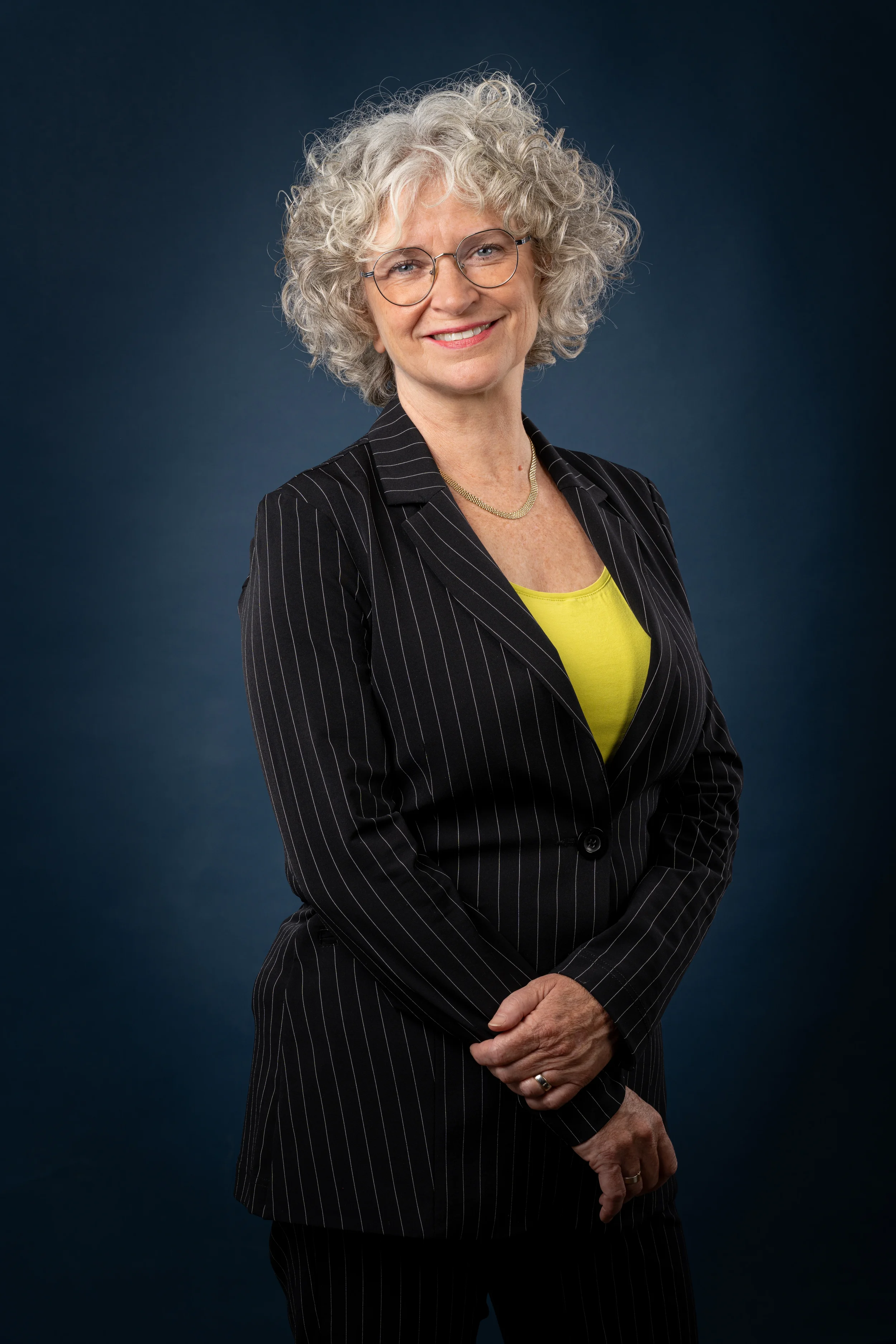 A woman with curly gray hair, wearing glasses, a black pinstripe suit, a yellow top, and a gold necklace, smiling against a dark blue background.