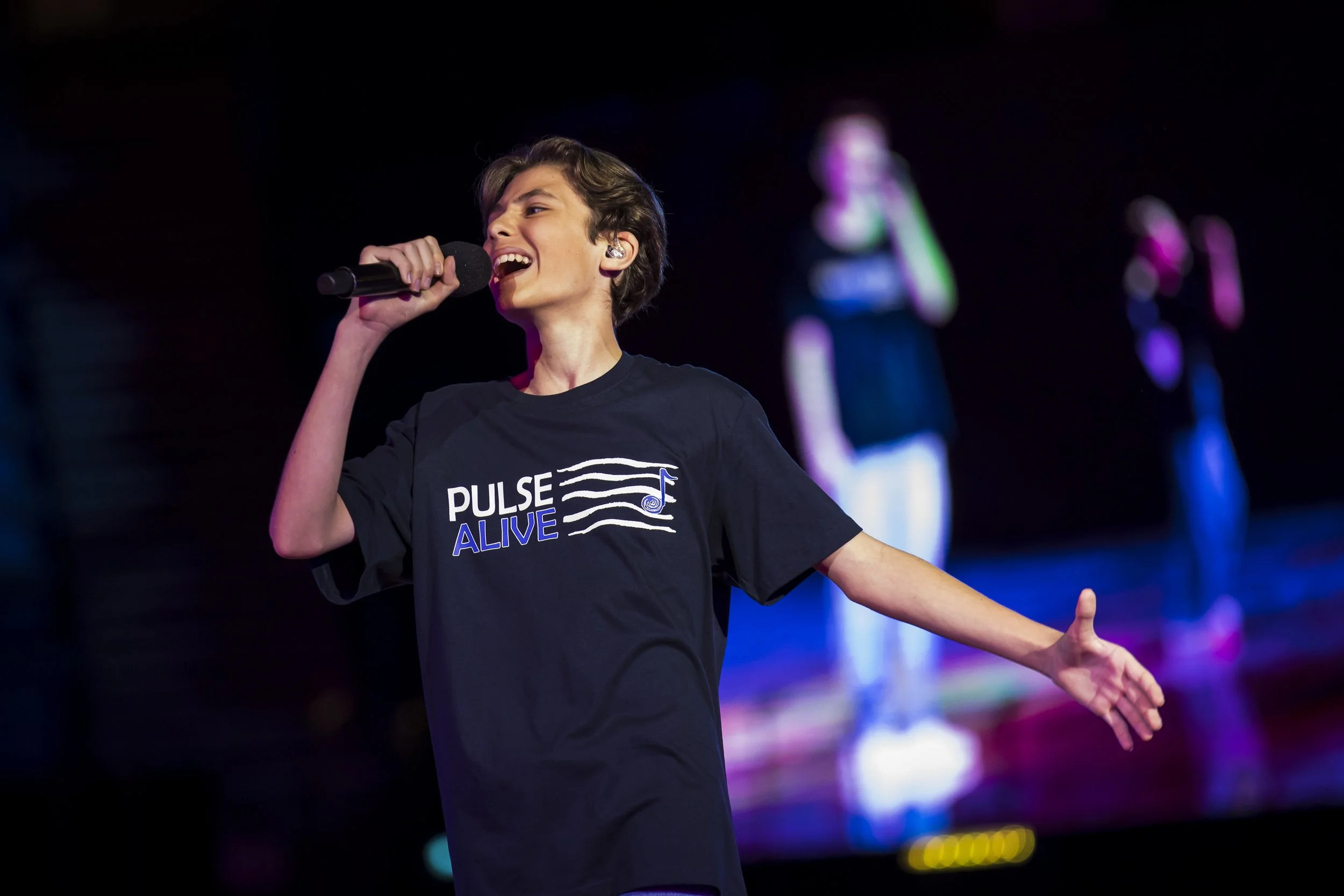 A young person singing into a microphone on stage with colorful lights in the background. They are wearing a black T-shirt with the words 'PULSE ALIVE' on it, and they have short, dark hair.