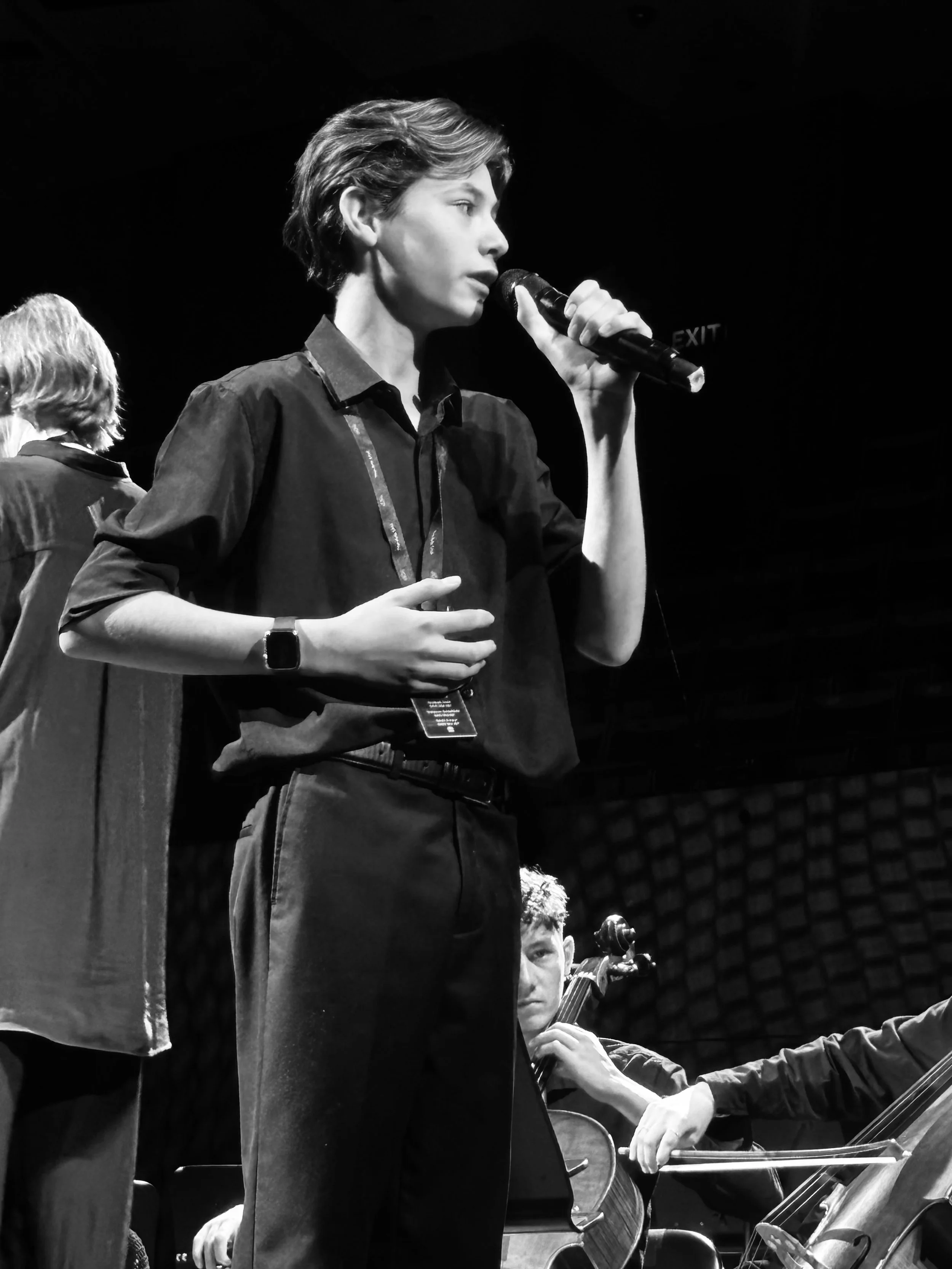 Black and white photo of a woman holding a microphone, singing or speaking on stage, with musicians and instruments in the background.