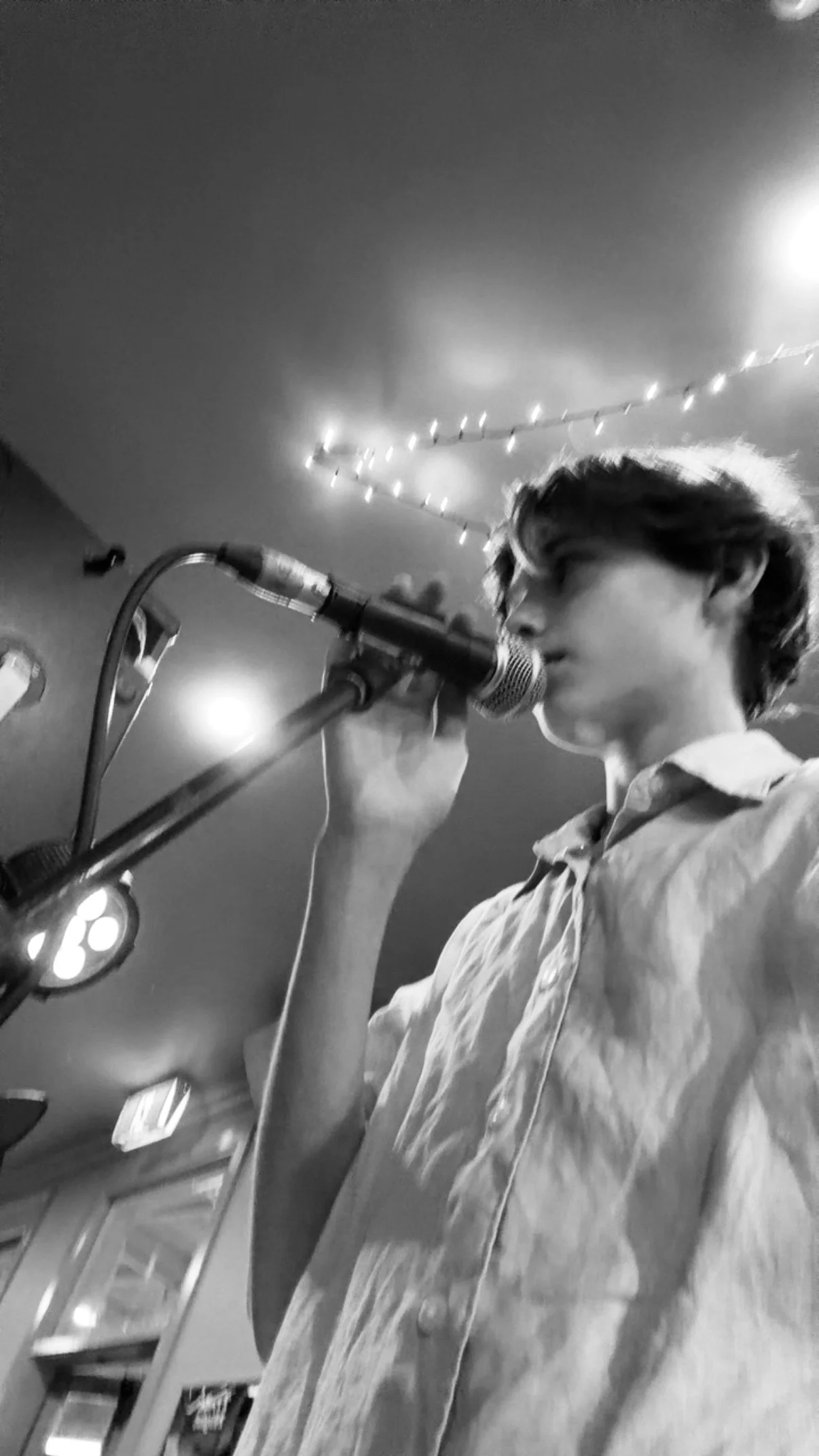 A person singing or speaking into a microphone in an indoor setting, with string lights hanging on the ceiling in the background.