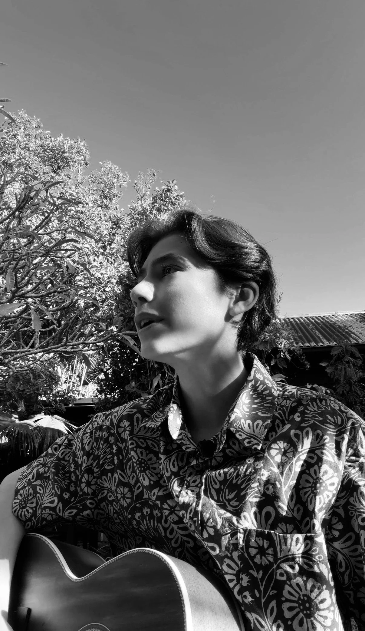 Black and white photo of a young person with medium-length hair wearing a patterned shirt, sitting outdoors with trees and a roof in the background, playing an acoustic guitar.