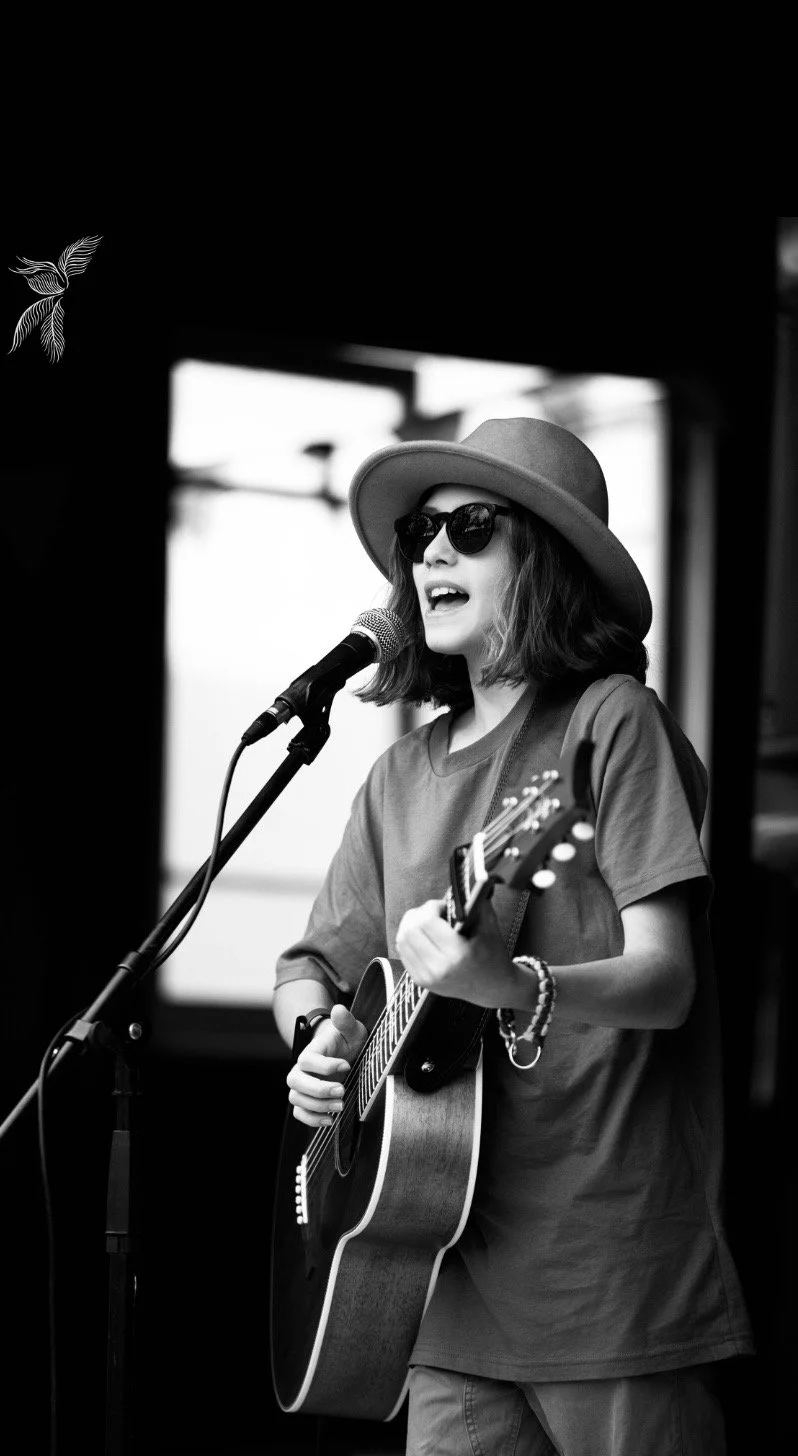 A woman wearing a hat and sunglasses singing into a microphone while playing an acoustic guitar.