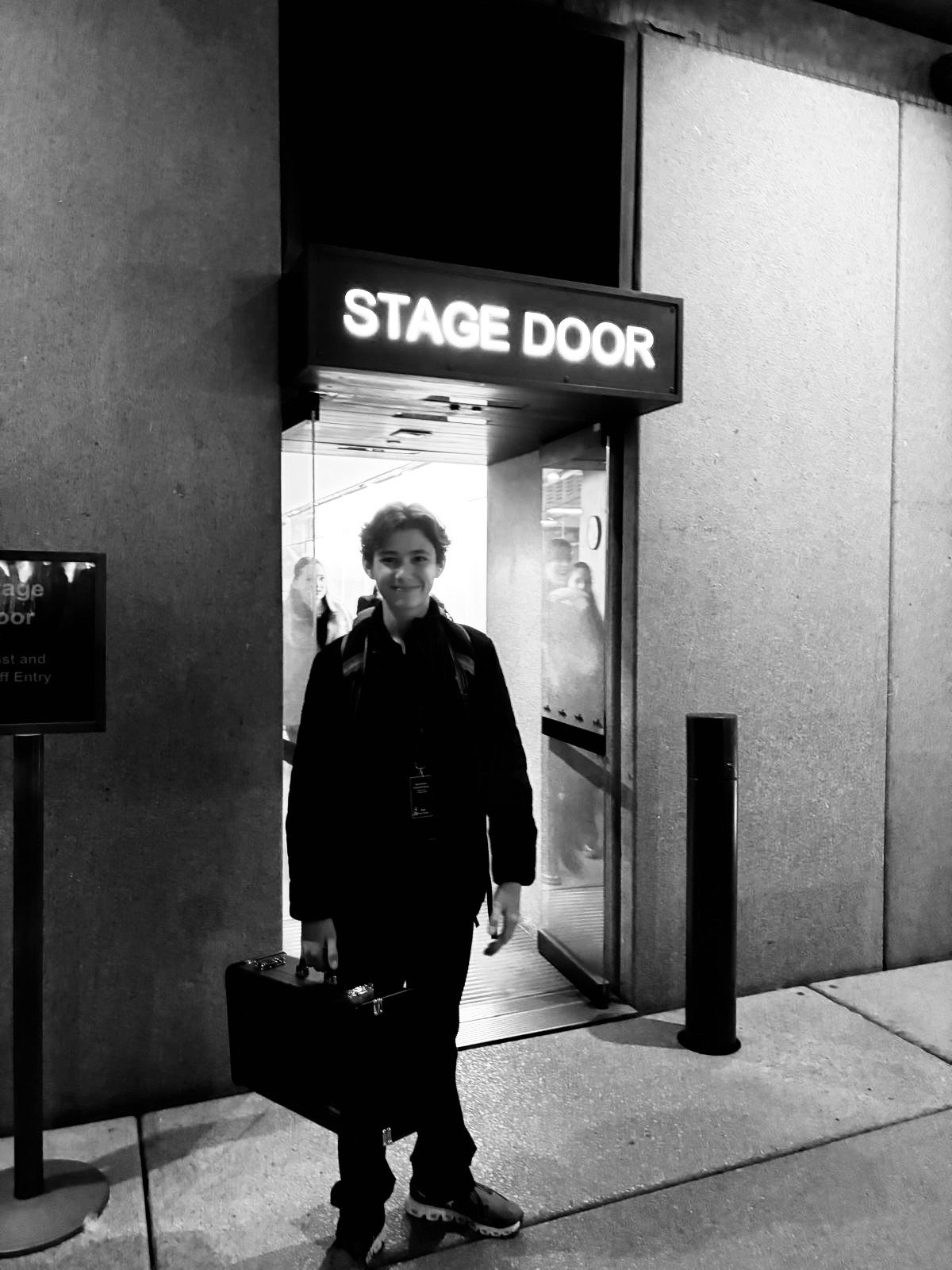A young boy standing outside a theater entrance with a sign that reads 'Stage Door'. He is smiling, dressed in dark clothing, and holding a suitcase.