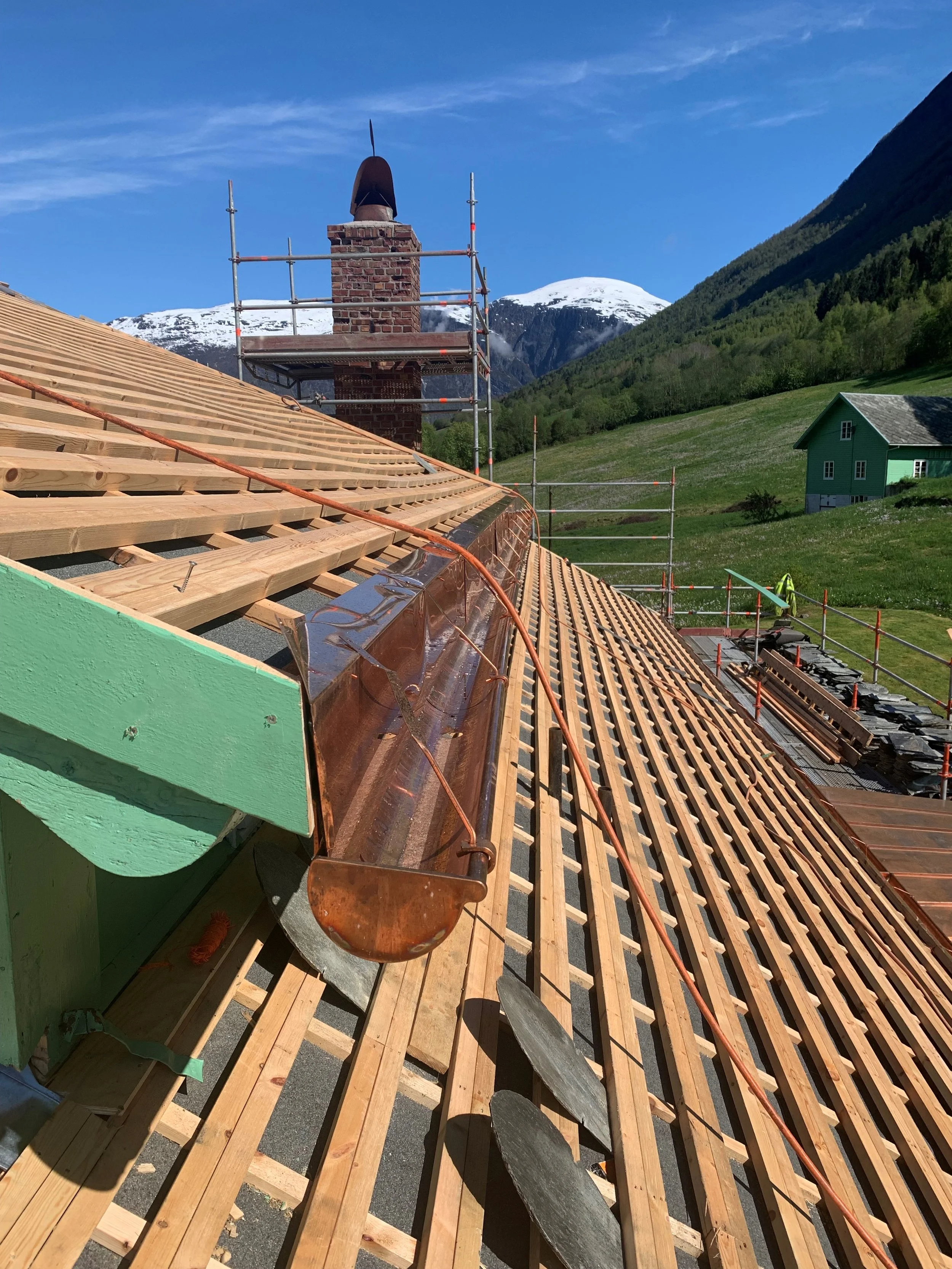 Takrenne under installasjon på et tak i en fjellregion med snødekte fjell i bakgrunnen og grønne åser og hytter i forgrunnen. Taket er i ferd med å bli dekket med nye takstein.