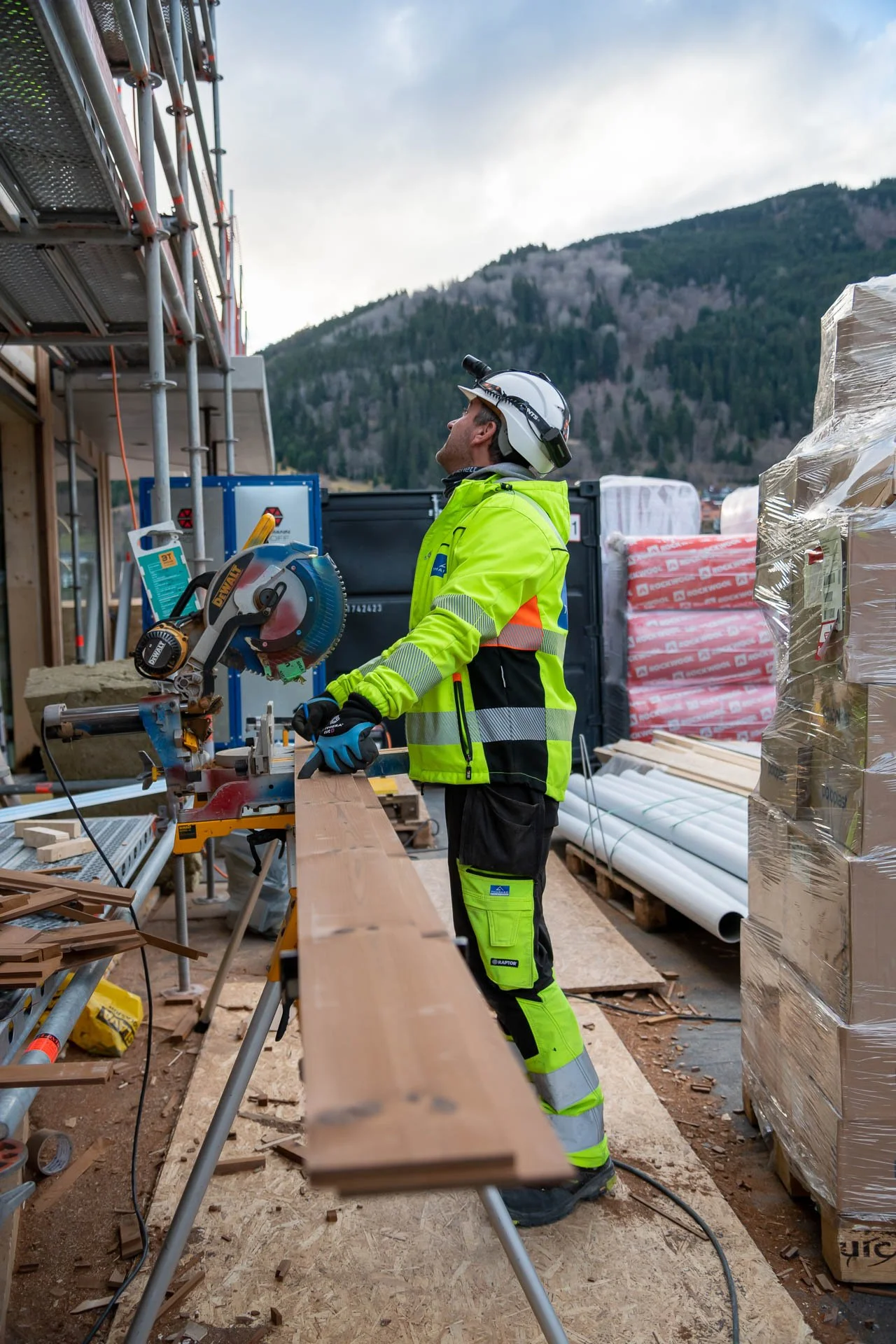 En bygningsarbeider i høyviskleding bruker en sirkelsag til å kutte treverk på en byggeplass med utstyr, paller og byggematerialer rundt seg, og med fjell og skog i bakgrunnen.
