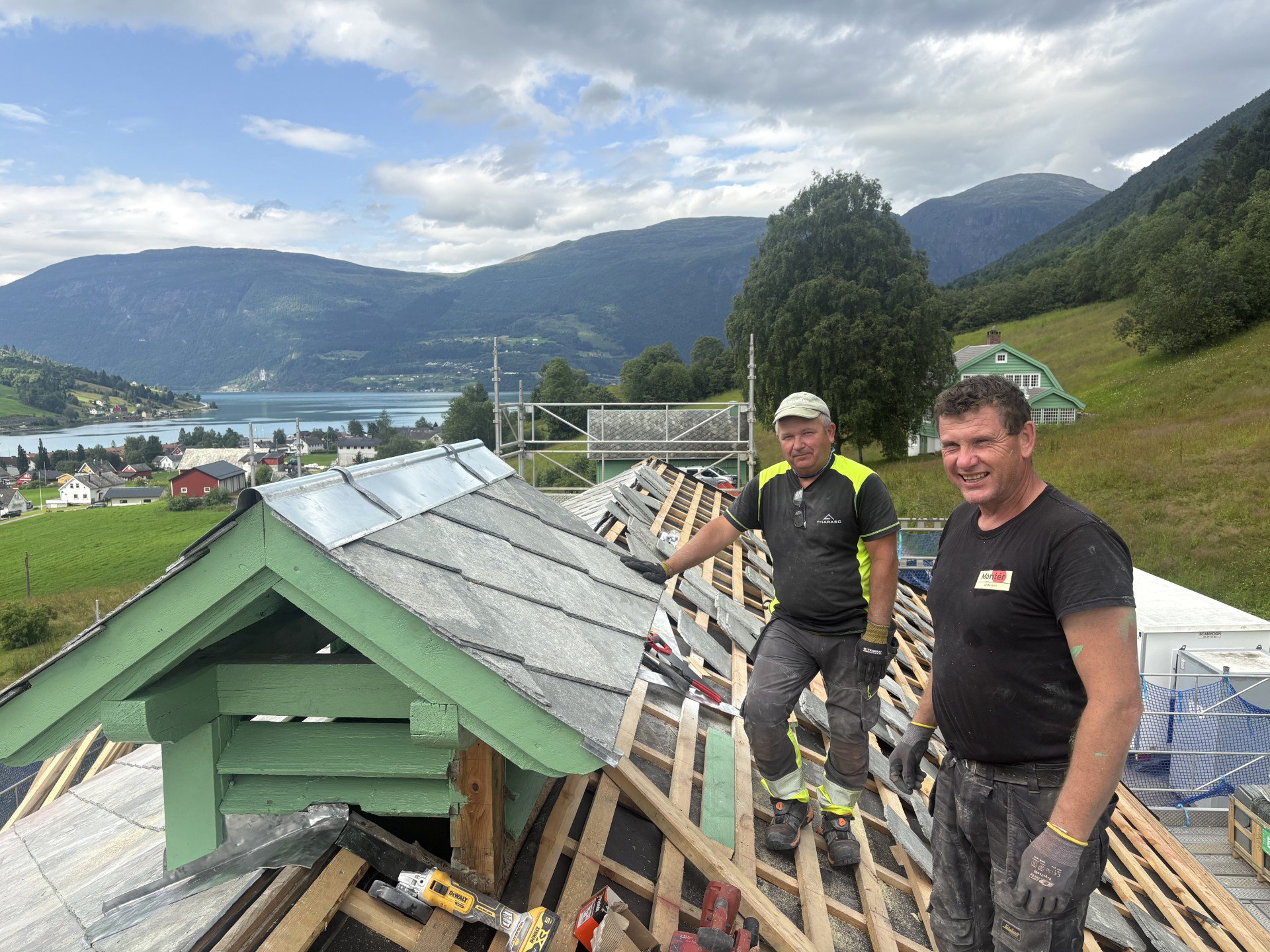 To menn står på et tak under konstruksjon av et hus. Bak dem er det et landskap med innsjø, hus og fjell, overskyet himmel med solglimt.