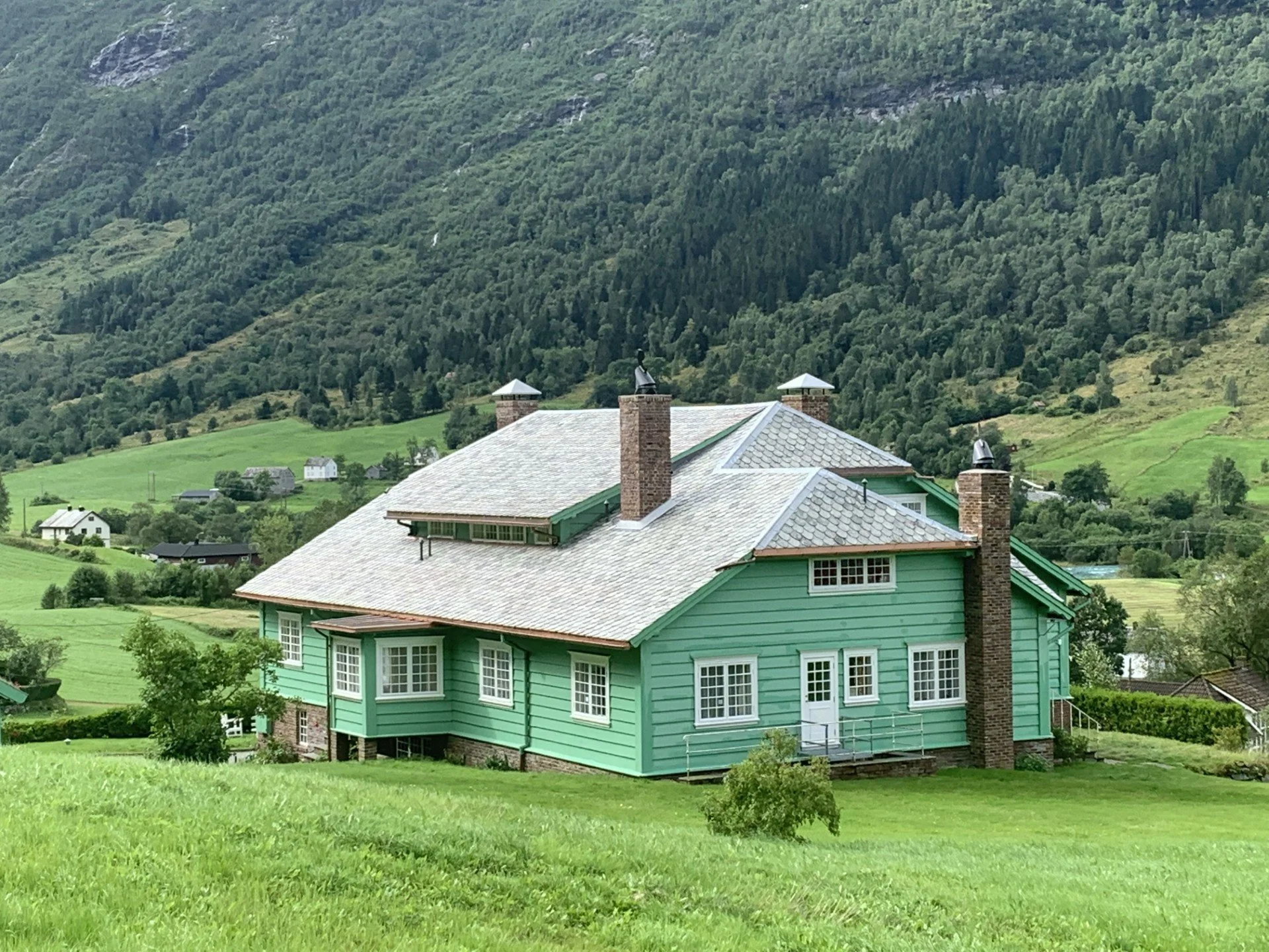Et grønt hus med hvite vinduer og skorstein, omgitt av grønne åser og fjell. Landskapet er kupert med flere små hus i bakgrunnen.