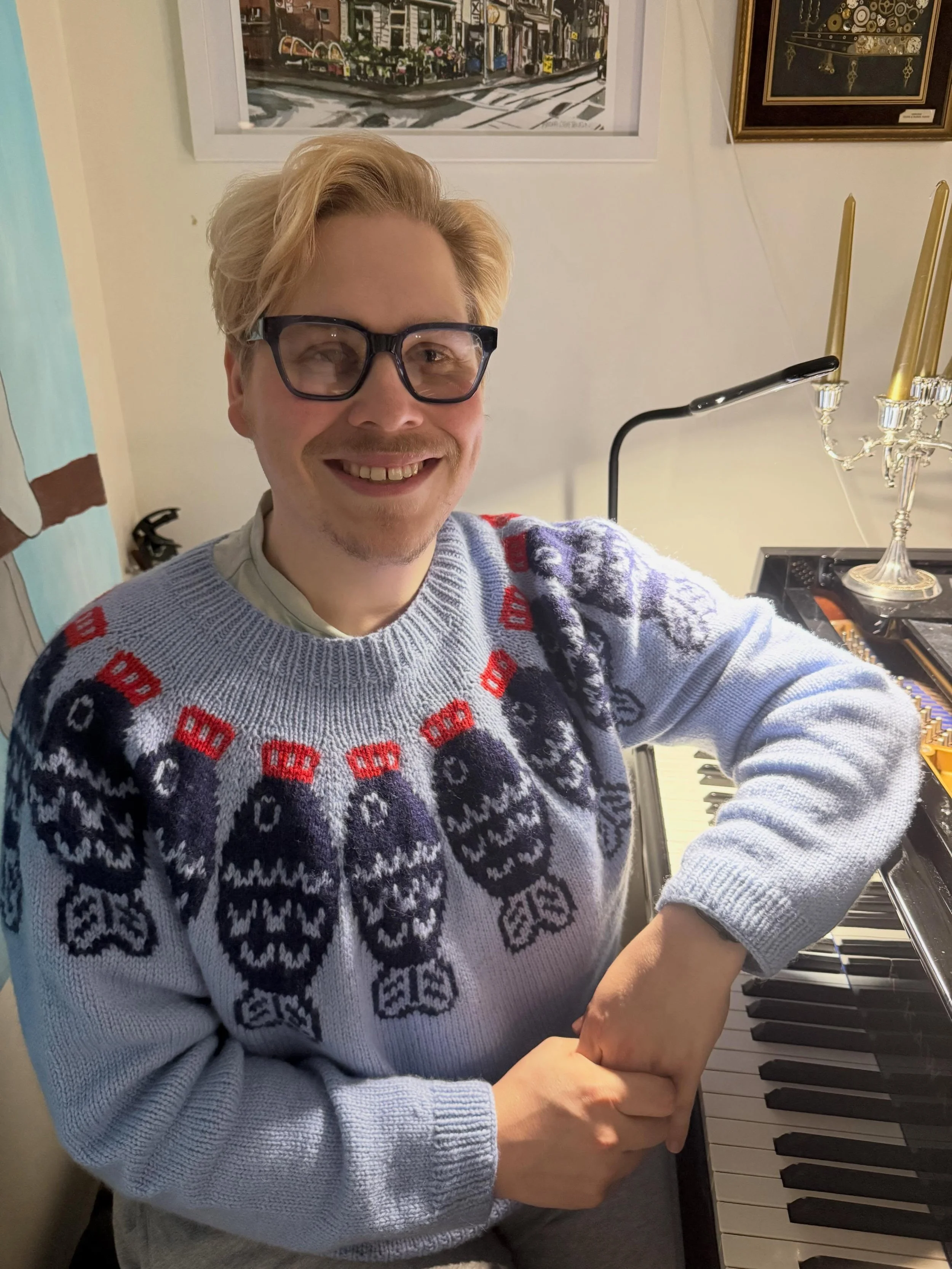 Photo of Kane Brown: He is a blonde man in dark blue glasses. He is wearing a light blue knitted jumper with soy sauce fish on it. He is sitting at a piano.