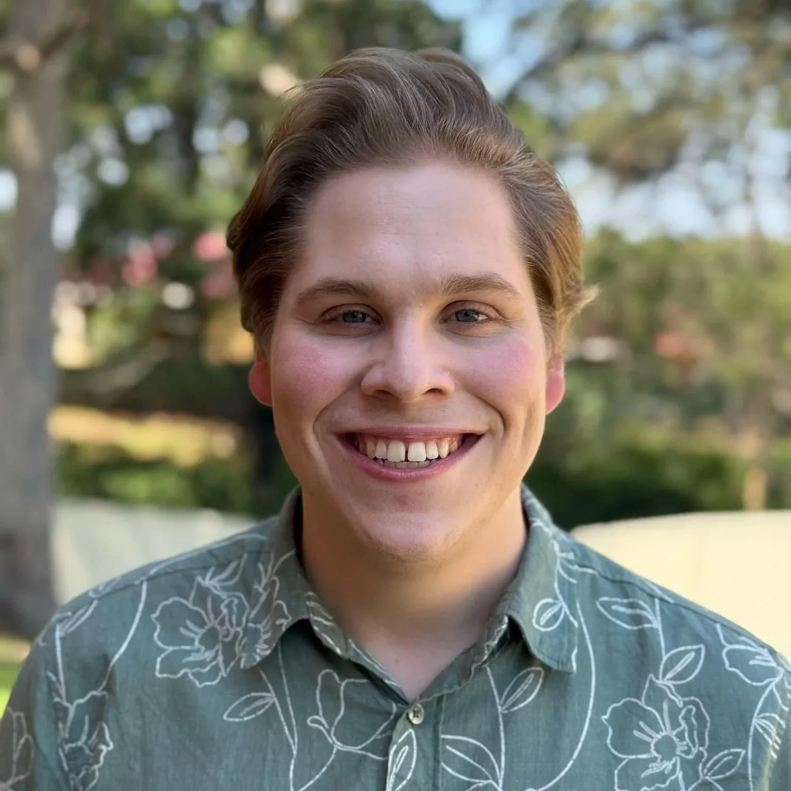 Photo of Kane Brown: He is a blonde man and is smiling. He is standing outside on a sunny day. He is wearing a green button up shirt with flowers on it.