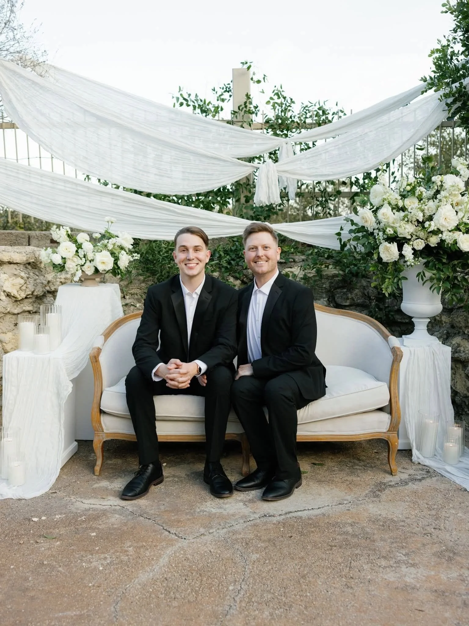 Two men dressed in black suits sitting on a white vintage sofa outdoors, with white drapes and flower arrangements behind them.