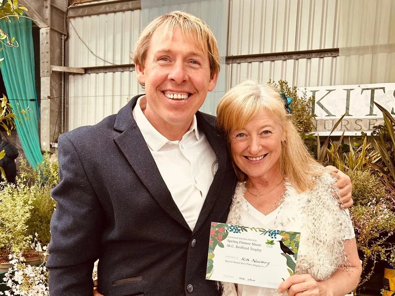 Two smiling people, a man in a suit and a woman holding a certificate, standing among plants inside a greenhouse or garden center.