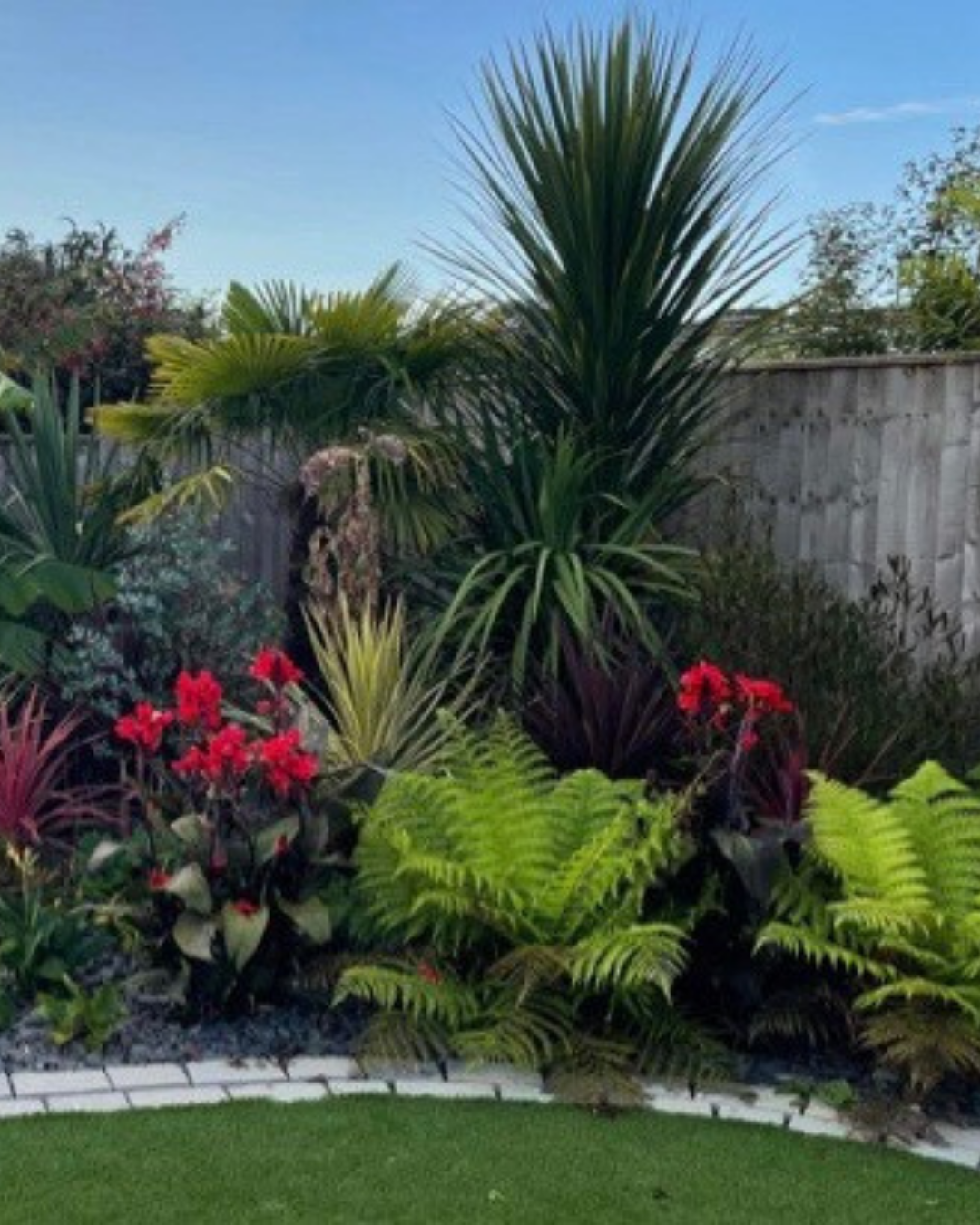 Lush tropical garden with various green plants, including large palm-like trees, colorful red flowers, and a manicured grassy area bordered by white bricks, with a wooden fence and clear blue sky in the background.