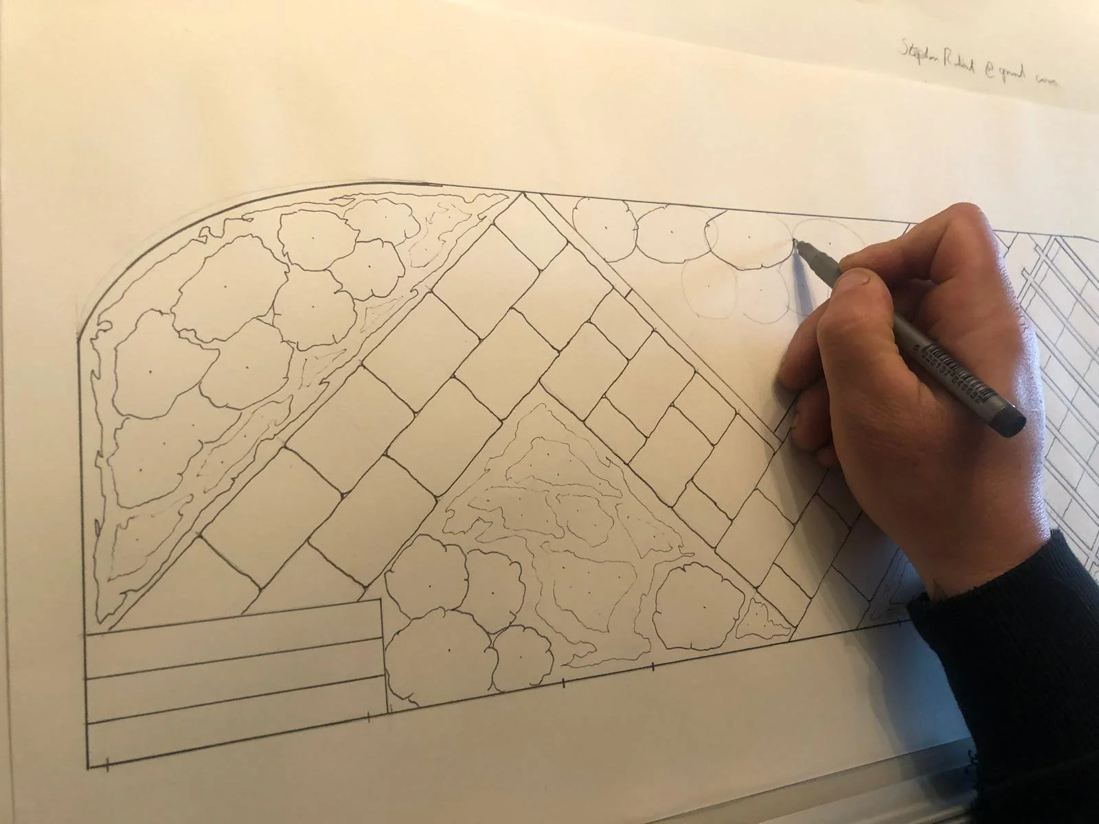 A person drawing a landscape plan on paper, featuring trees, pathways, and landscaped areas.