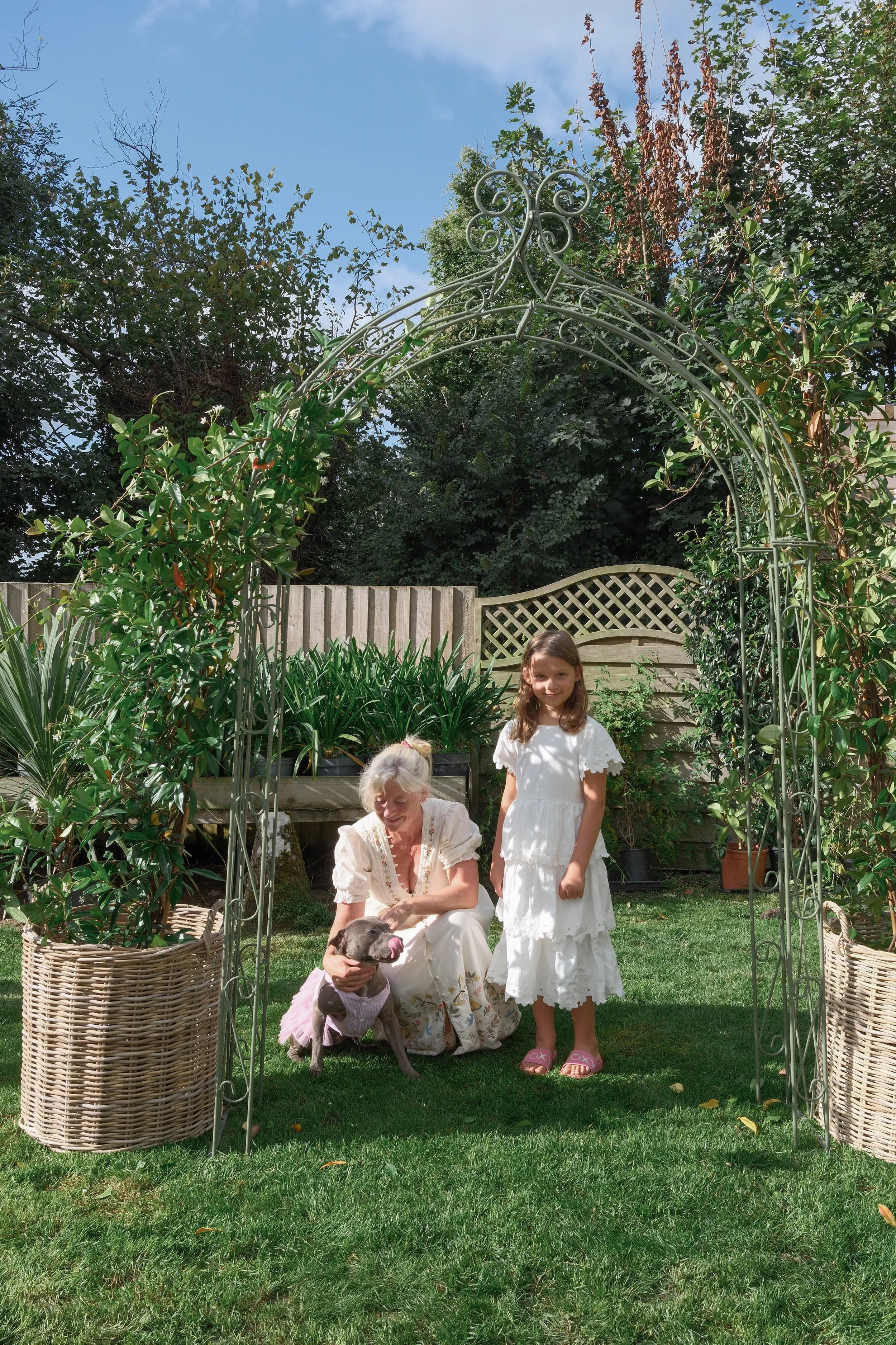 A woman with blonde hair in a light-colored dress sitting on the grass petting a brown and white dog wearing a pink tutu. Two girls in white dresses stand nearby under a decorative metal archway in a lush garden.