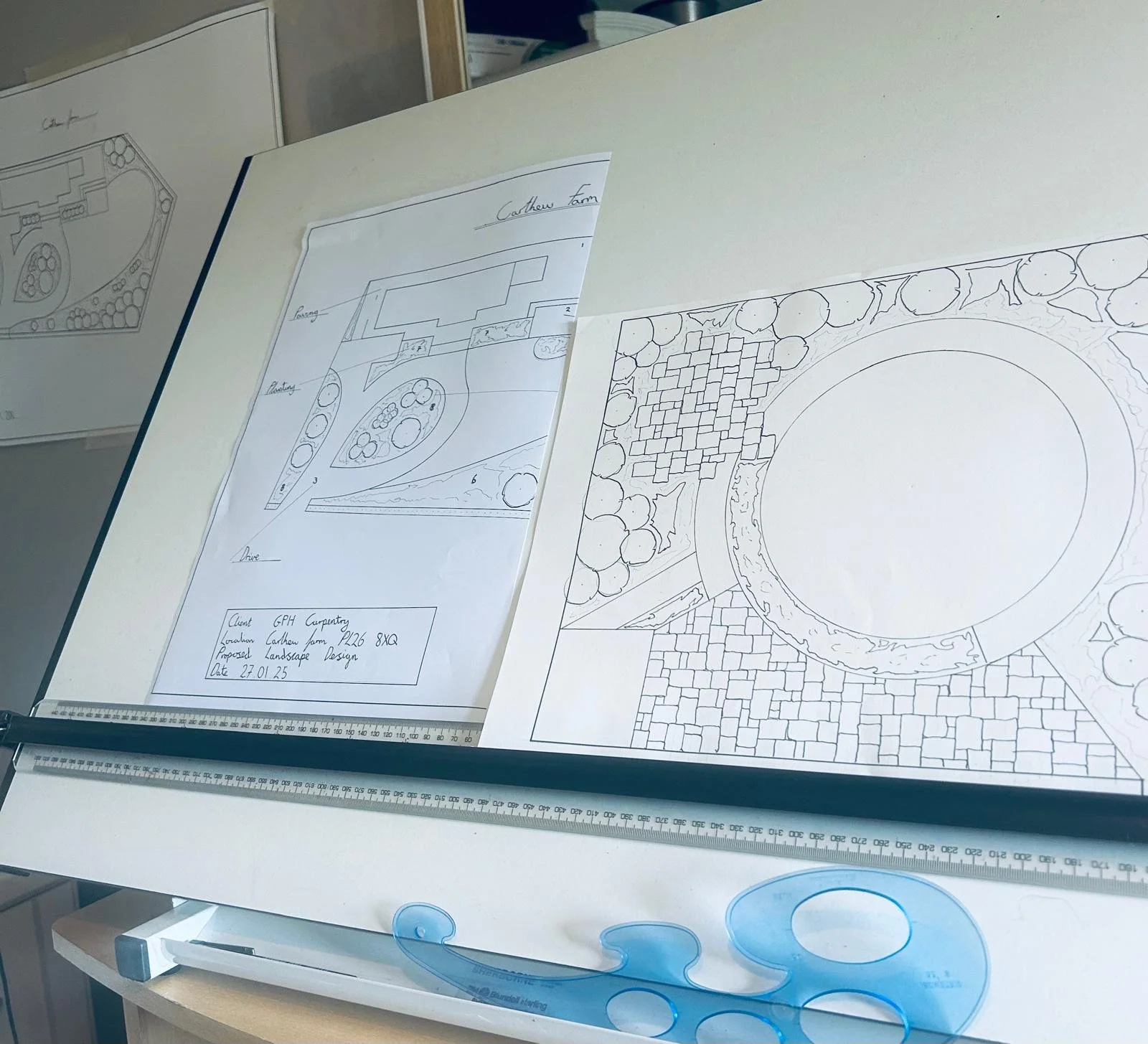 Design sketches and plans for landscape architecture, including layout and plant placement, on large sheets of paper with a ruler and a blue drafting tool on the table.