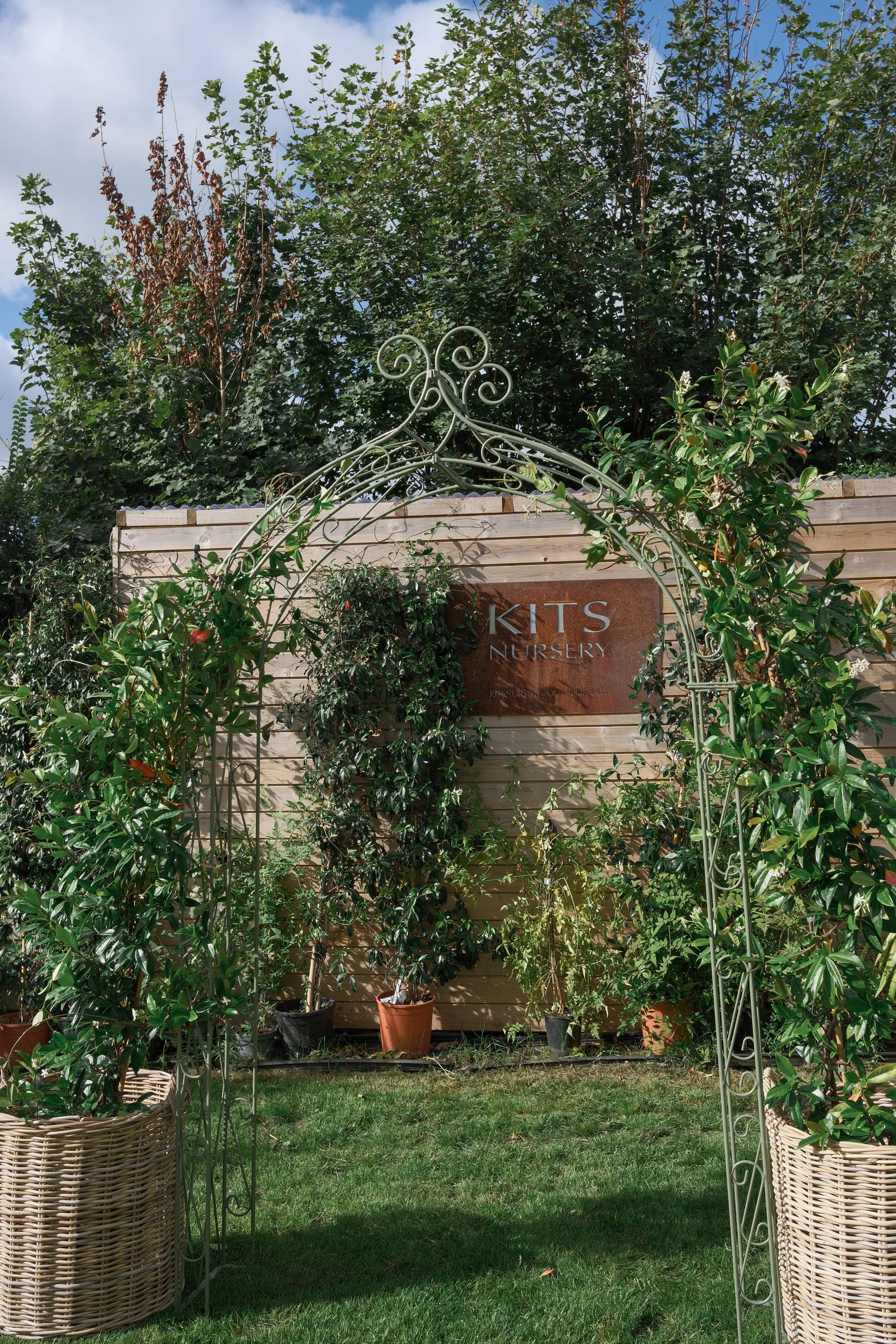 A garden scene with potted plants and a decorative garden arch in front of a wooden fence. A sign reading 'KITS Nursery' is attached to the fence. Tall trees with green and brown leaves are in the background under a partly cloudy sky.