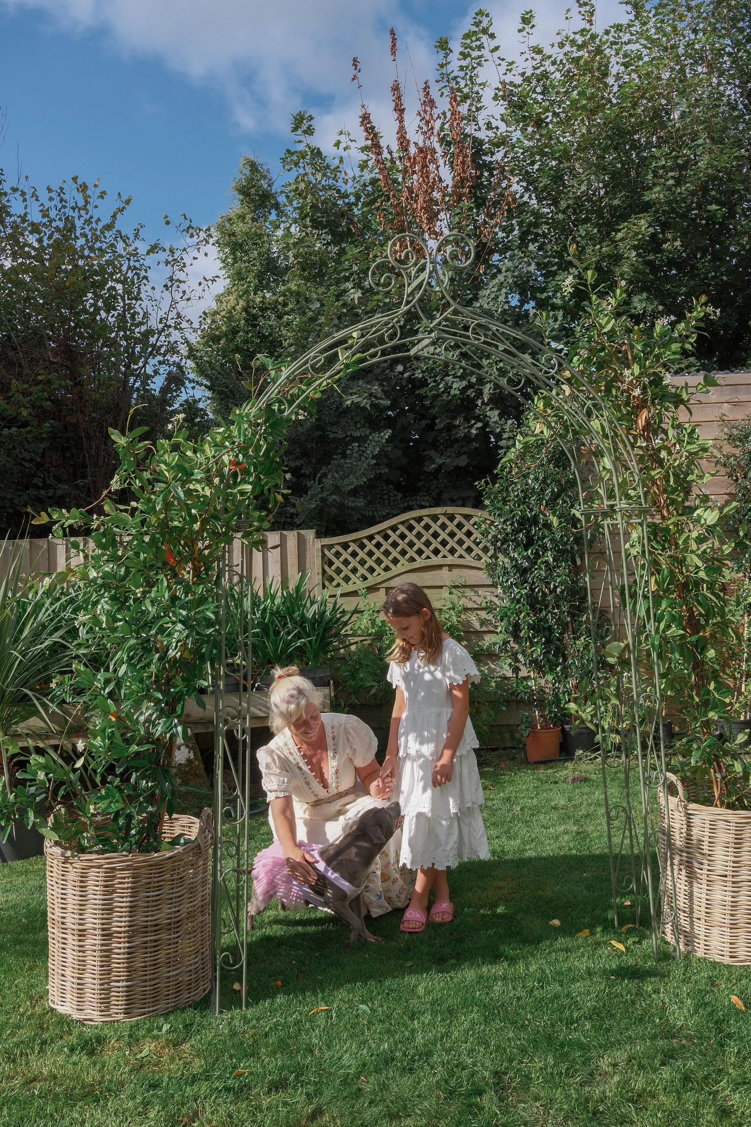 A woman and two girls standing under a garden arch, with a dog wearing a tutu, in a backyard with green grass, plants, and trees. Two wicker baskets are on either side of the arch.